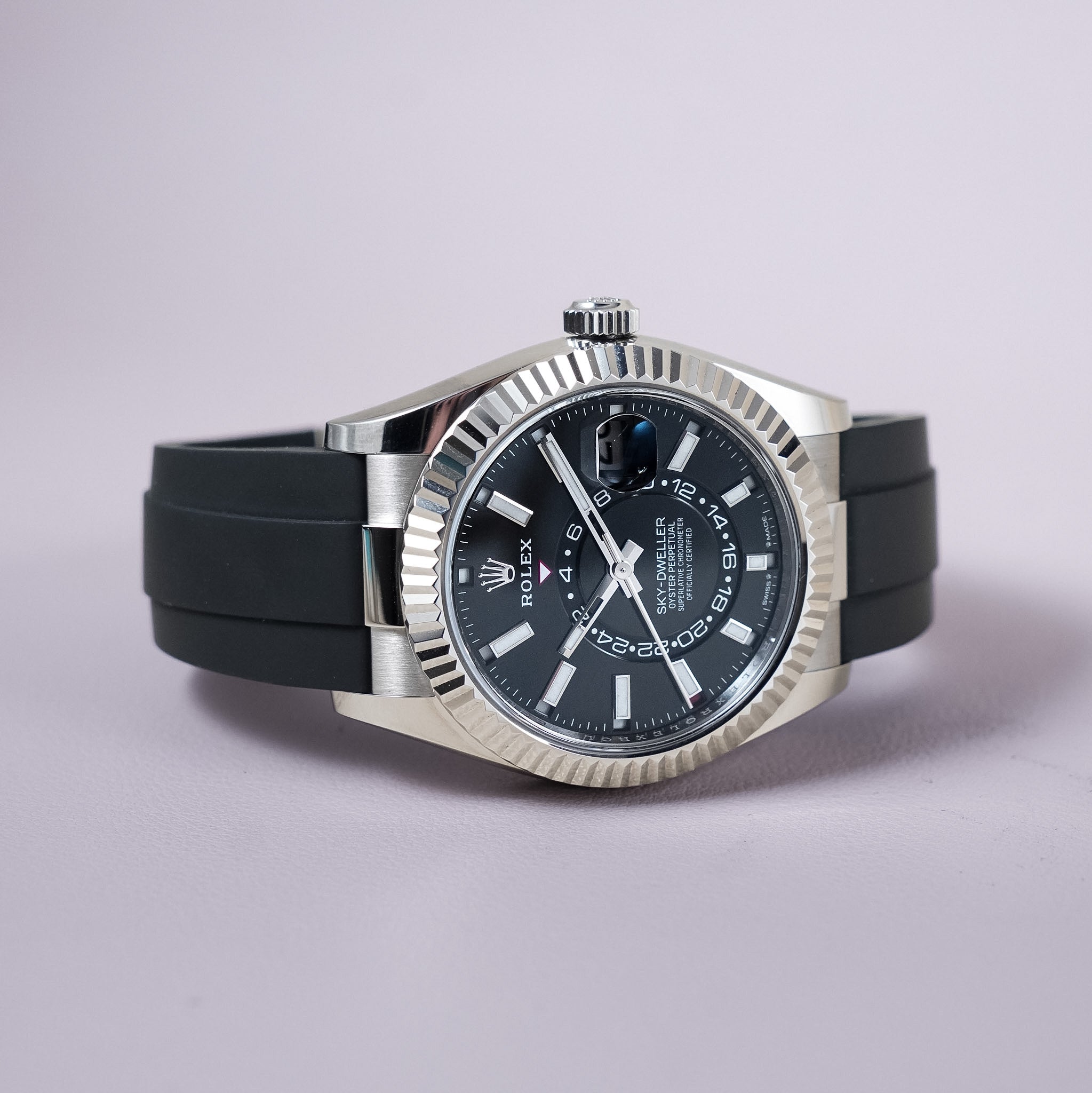 Rolex Sky-Dweller wristwatch with a white gold fluted bezel, black dial, and black rubber strap, photographed on a light lavender background.