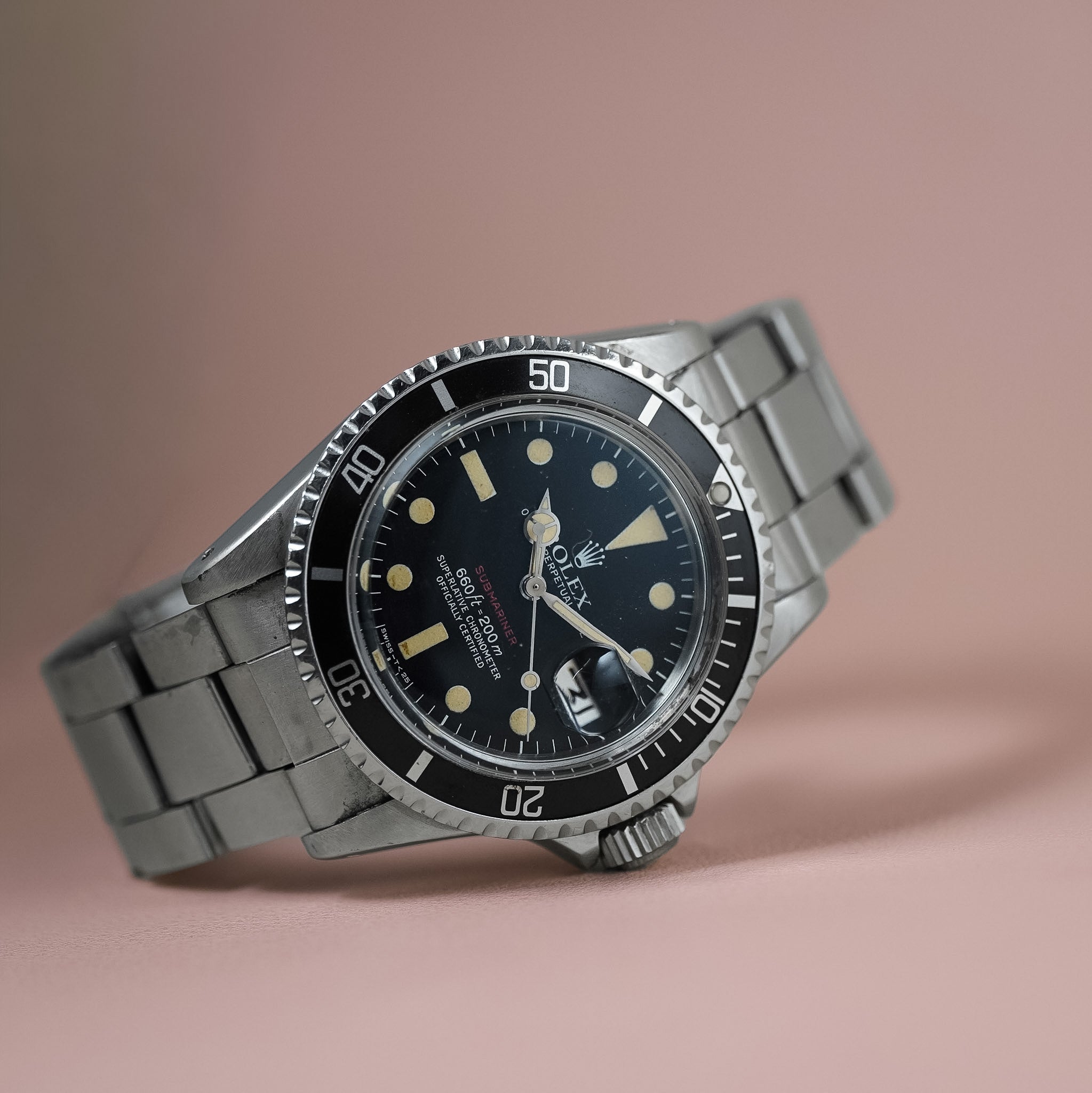 A vintage Rolex Submariner watch with a black dial, black bezel, and a silver-toned stainless steel bracelet, resting on a solid dusty pink background.