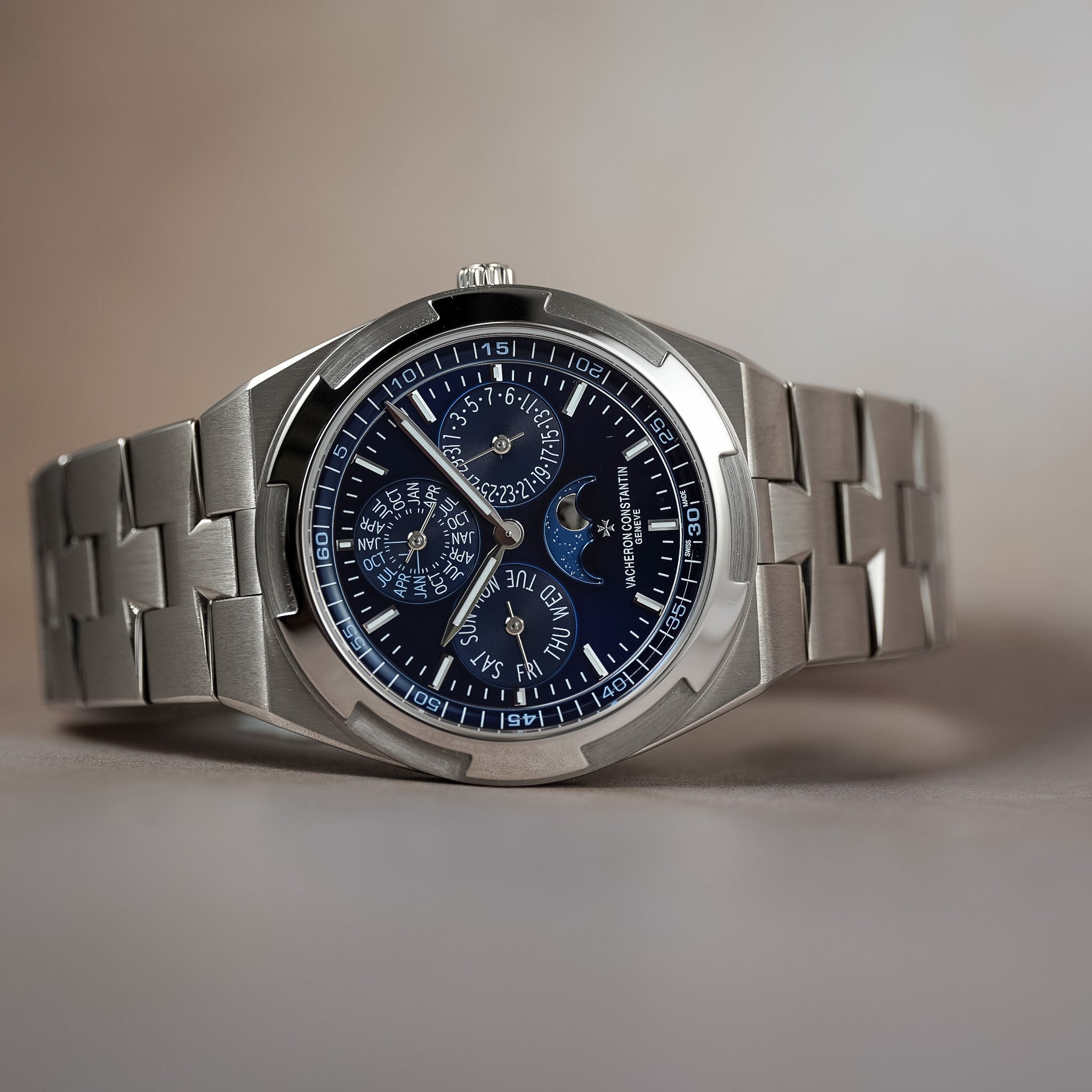 A luxury stainless steel wristwatch with a silver-tone metal bracelet and a dark blue dial featuring multiple subdials, including a moon phase indicator, photographed on a neutral beige background.
