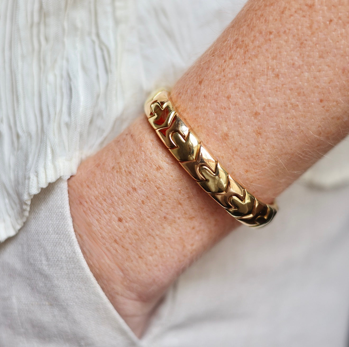 A close-up of a person's wrist wearing a polished gold chain-link bangle bracelet, with a white textured top in the background.