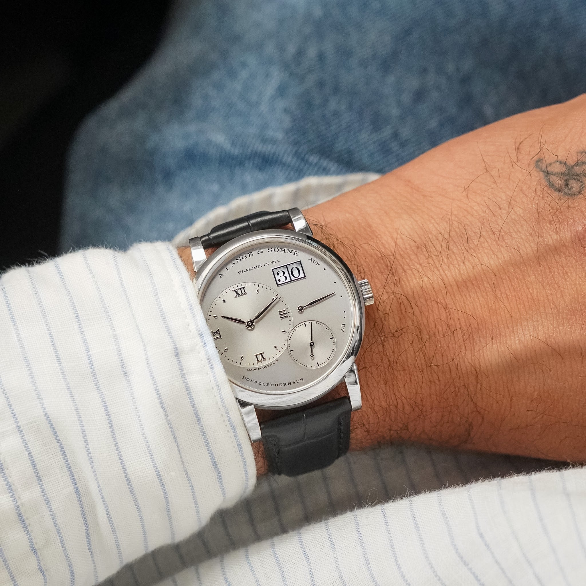 A close-up of a wrist wearing a silver-toned Perpetual watch with a black leather strap, a white dial with Roman numerals, and a day/date window. The person is wearing a white striped shirt and blue jeans.