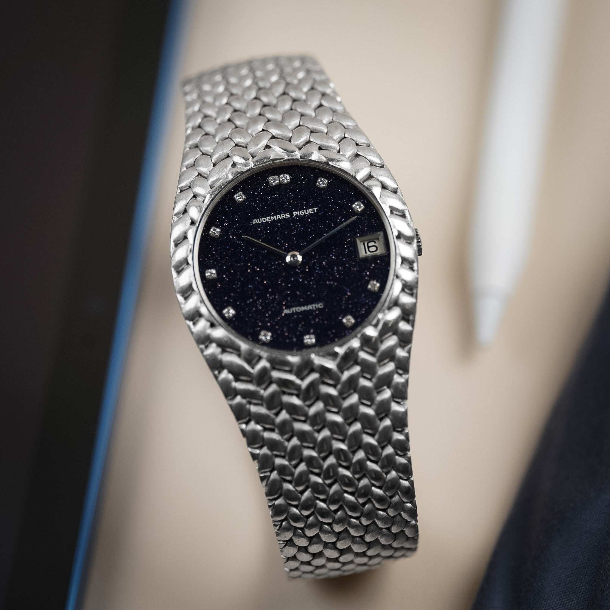 An Audemars Piguet wristwatch with a woven white gold case, diamond hour markers, date window, and a sparkly blue aventurine dial photographed against a blurry background. 