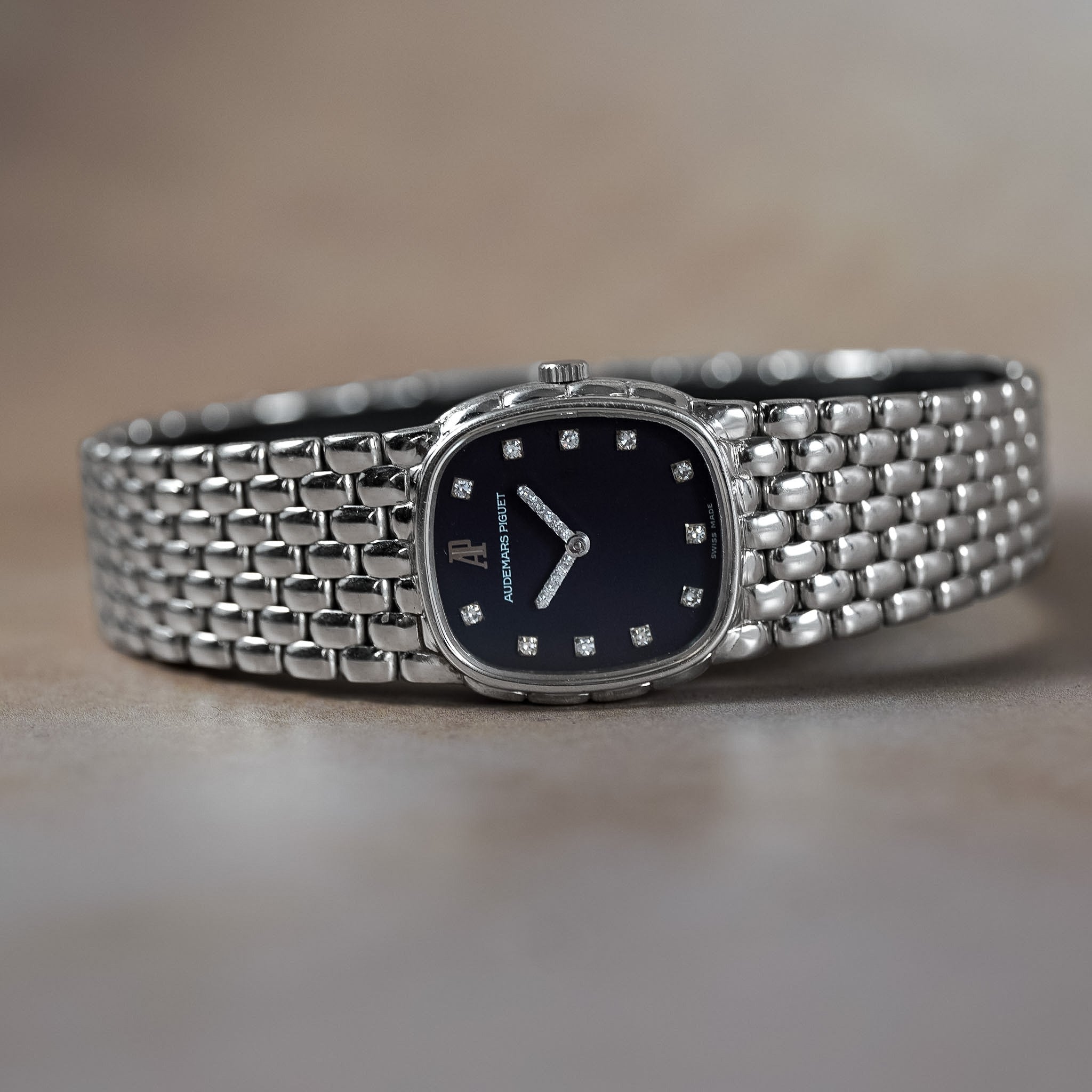 A close-up shot of a luxury watch with a silver-toned metal mesh bracelet and a dark navy blue dial featuring diamond hour markers.