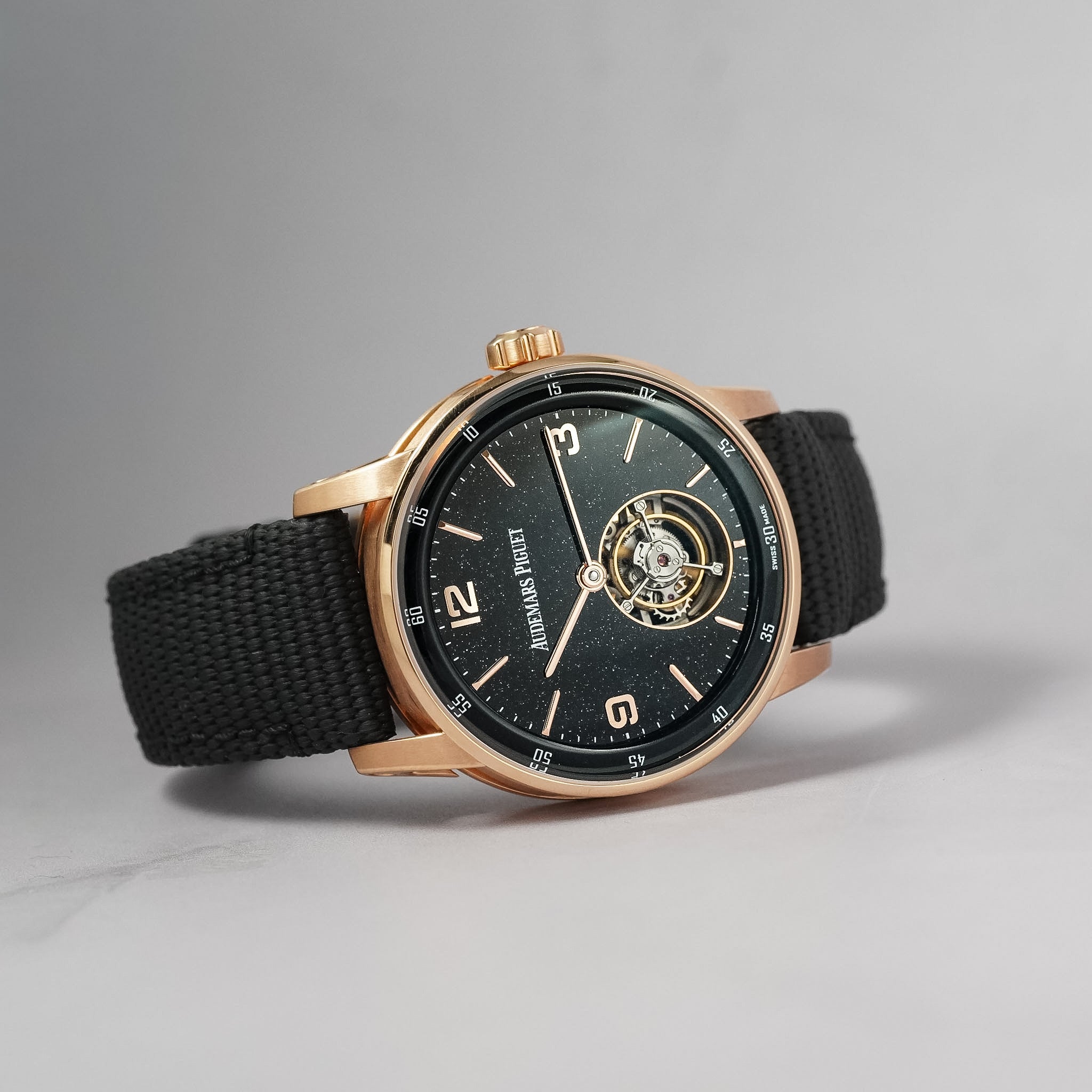 A wristwatch with a black fabric strap, a rose gold case, and a black dial featuring a visible automatic(tourbillon) movement through an openwork design. The brand name 'AUDEMARS PIGUET' is printed on the dial.