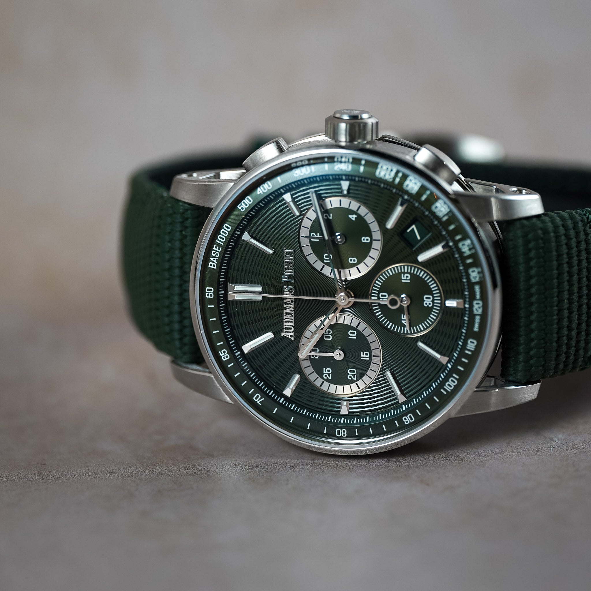 An Audemars Piguet chronograph watch with a dark green dial, silver-tone stainless steel case, and a dark green fabric strap, resting on a neutral gray surface.