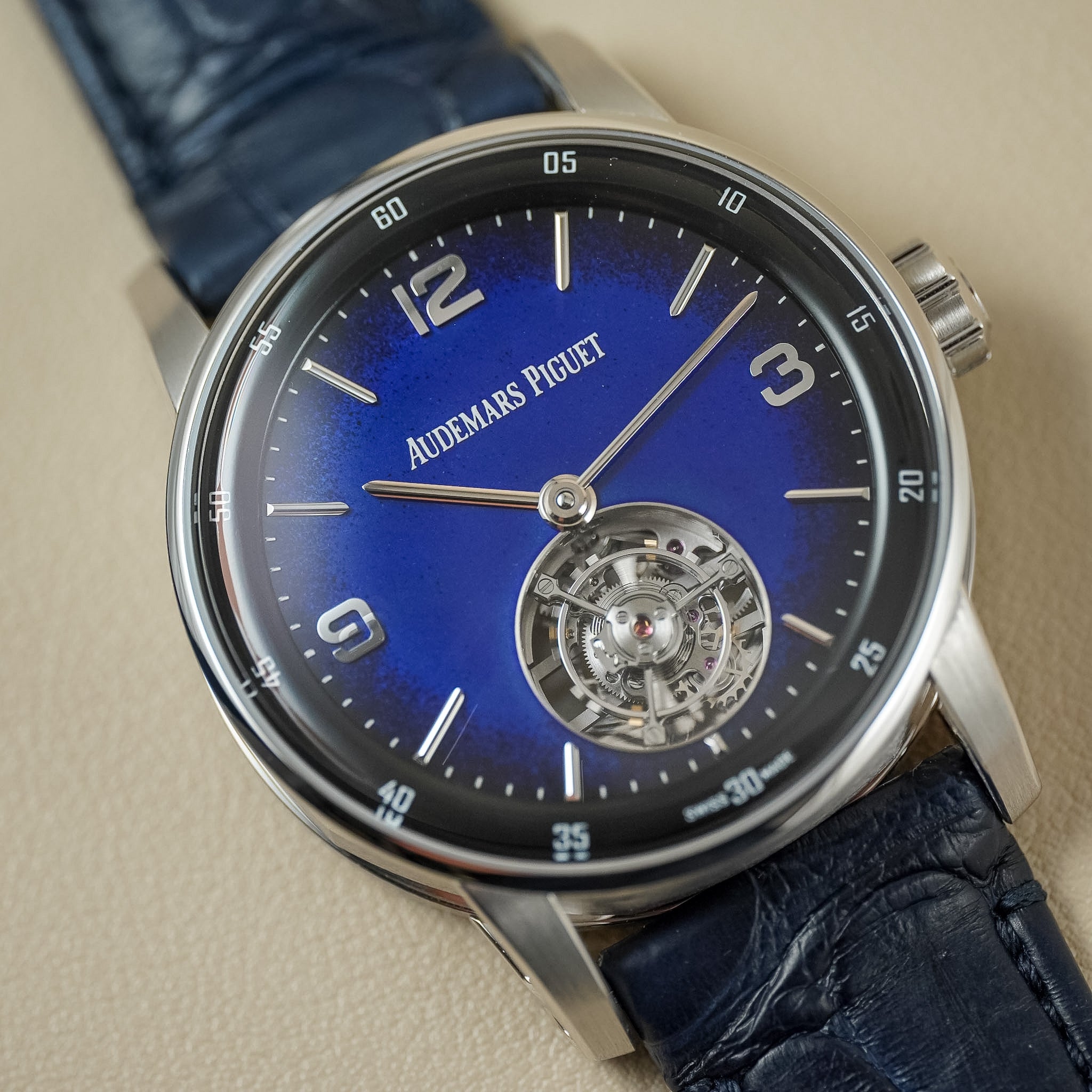 A close-up shot of an Audemars Piguet wristwatch featuring a vibrant blue sunburst dial, a visible tourbillon movement at the 6 o'clock position, and a dark blue alligator leather strap on a neutral beige background.
