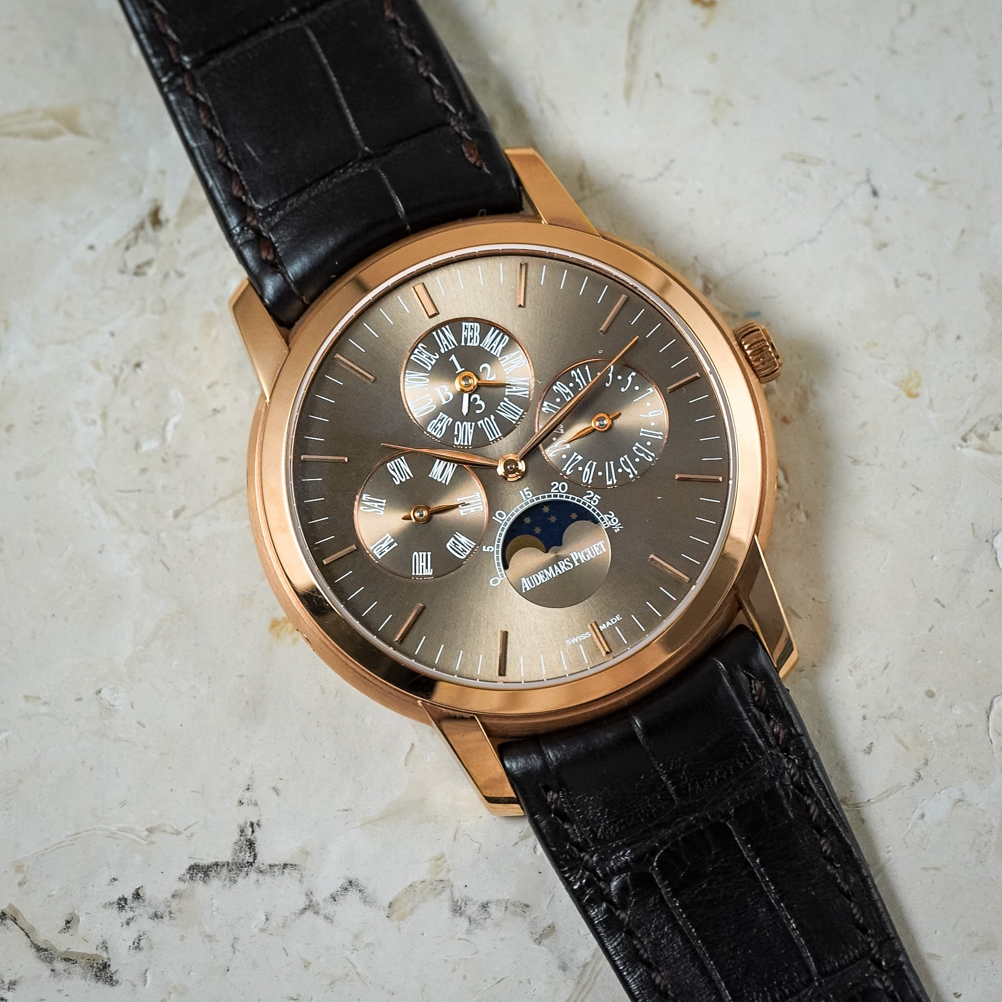 A luxury wristwatch with a rose gold case, a complex multi-dial grey dial, and a black alligator leather strap, resting on a textured light beige stone surface.