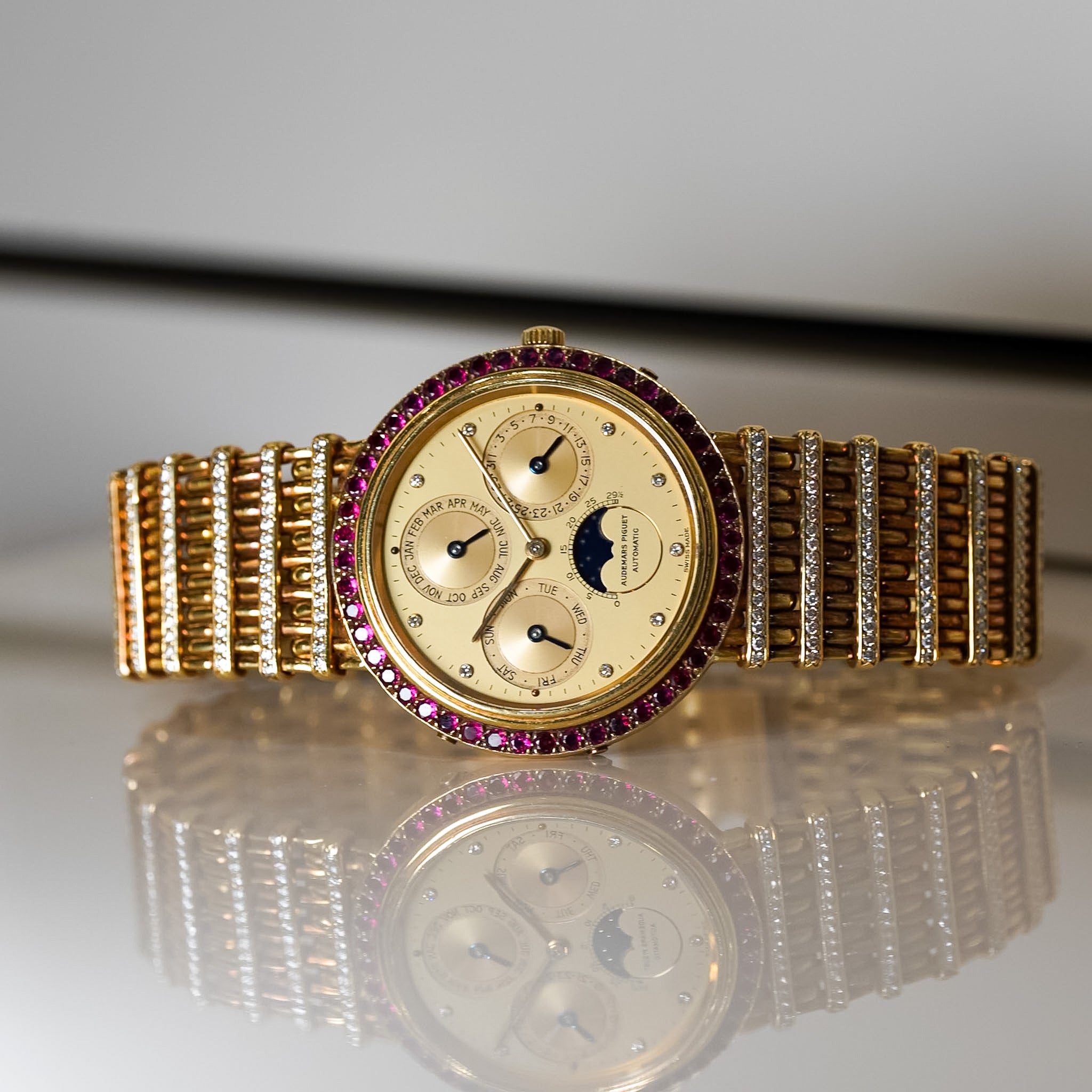 A yellow gold perpetual calendar watch laying sideways on a white reflective table. The watch is accented with rubies and diamonds.