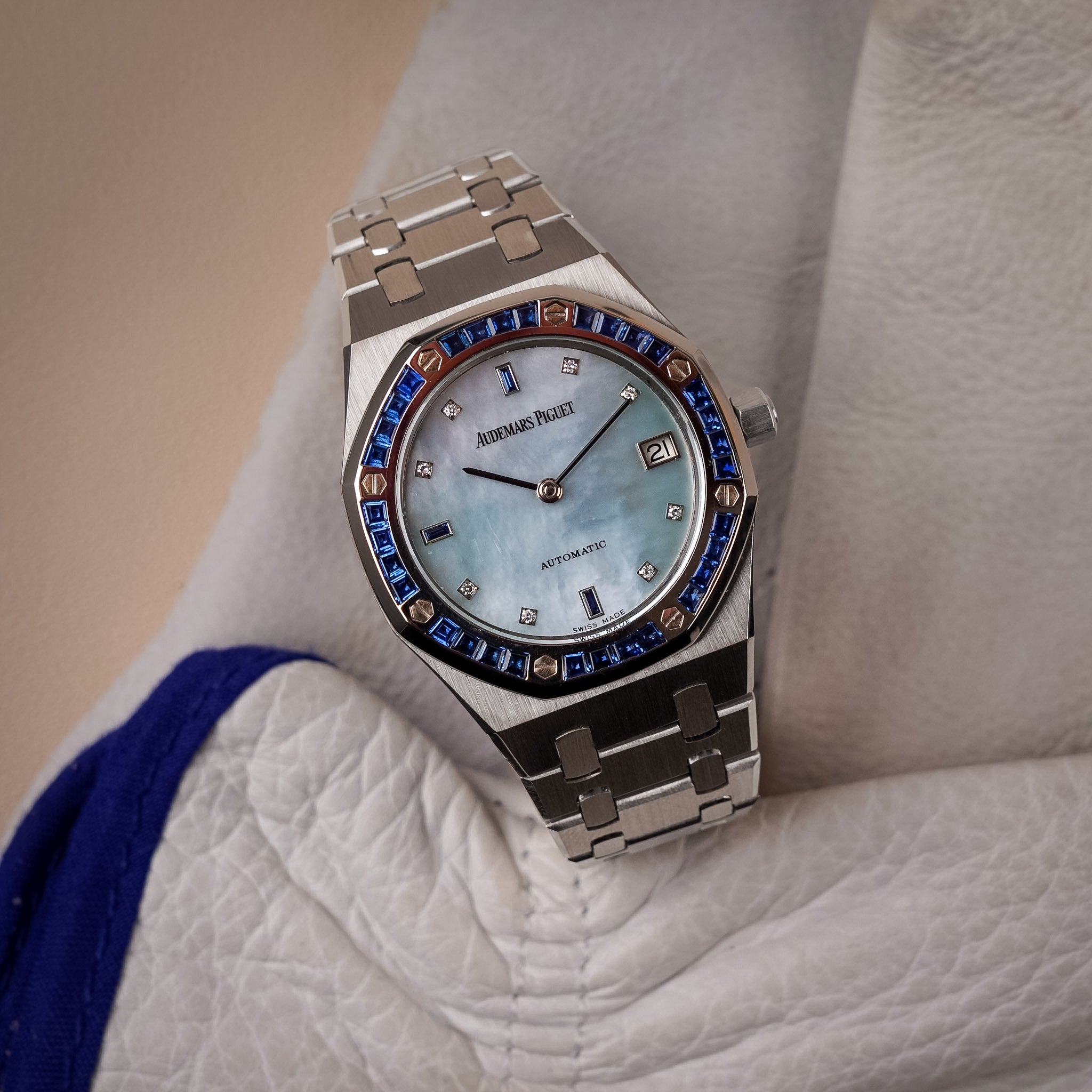 A white gold Audemars Piguet Royal Oak with a blue sapphire bezel, a blue mother of pearl dial, and blue sapphire and diamond hour markers. Shot at a slight angle sitting on white leather with blue trim.