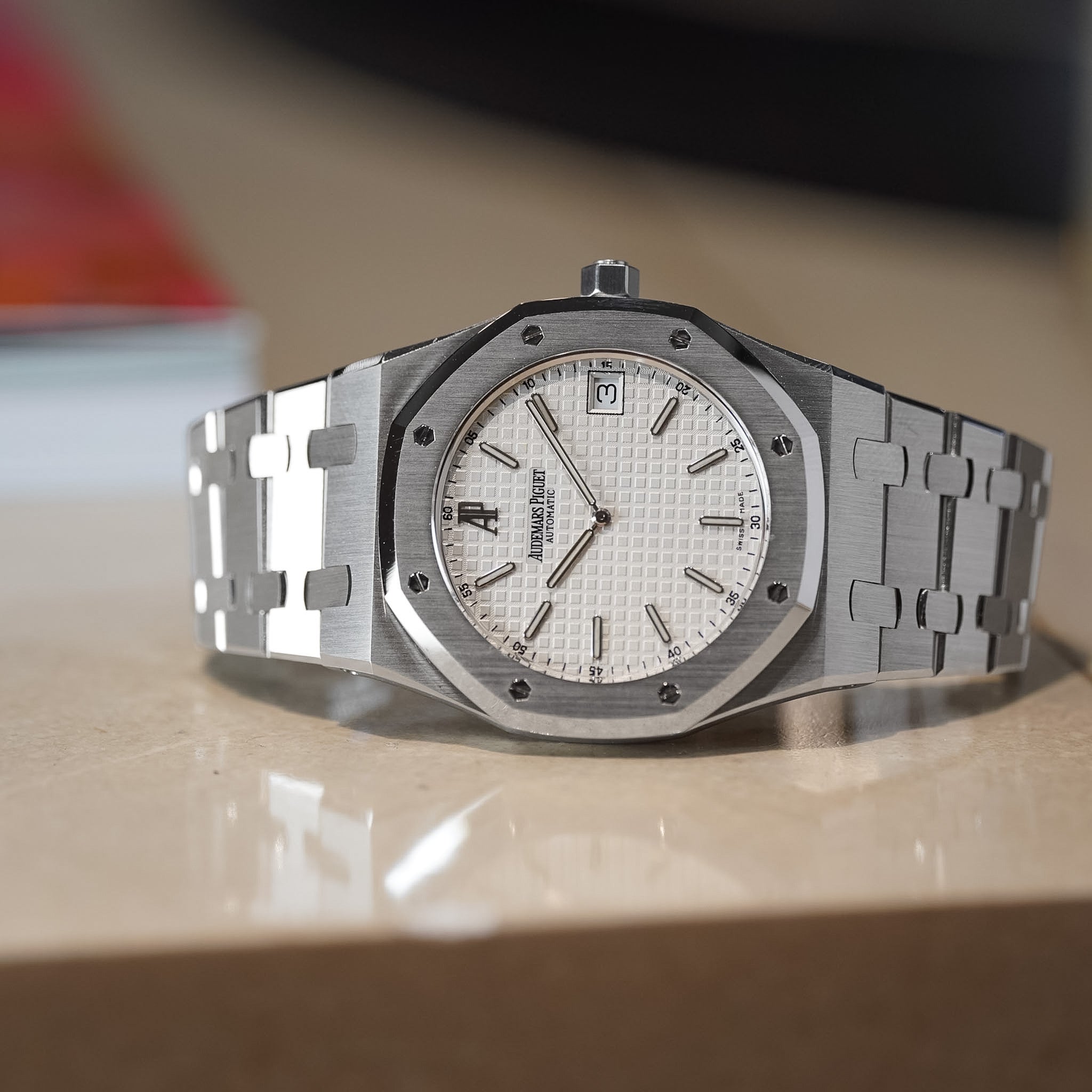 A stainless steel wristwatch with an octagonal bezel, silver-toned metal bracelet, and a white textured dial with a date window, resting on a reflective beige surface.