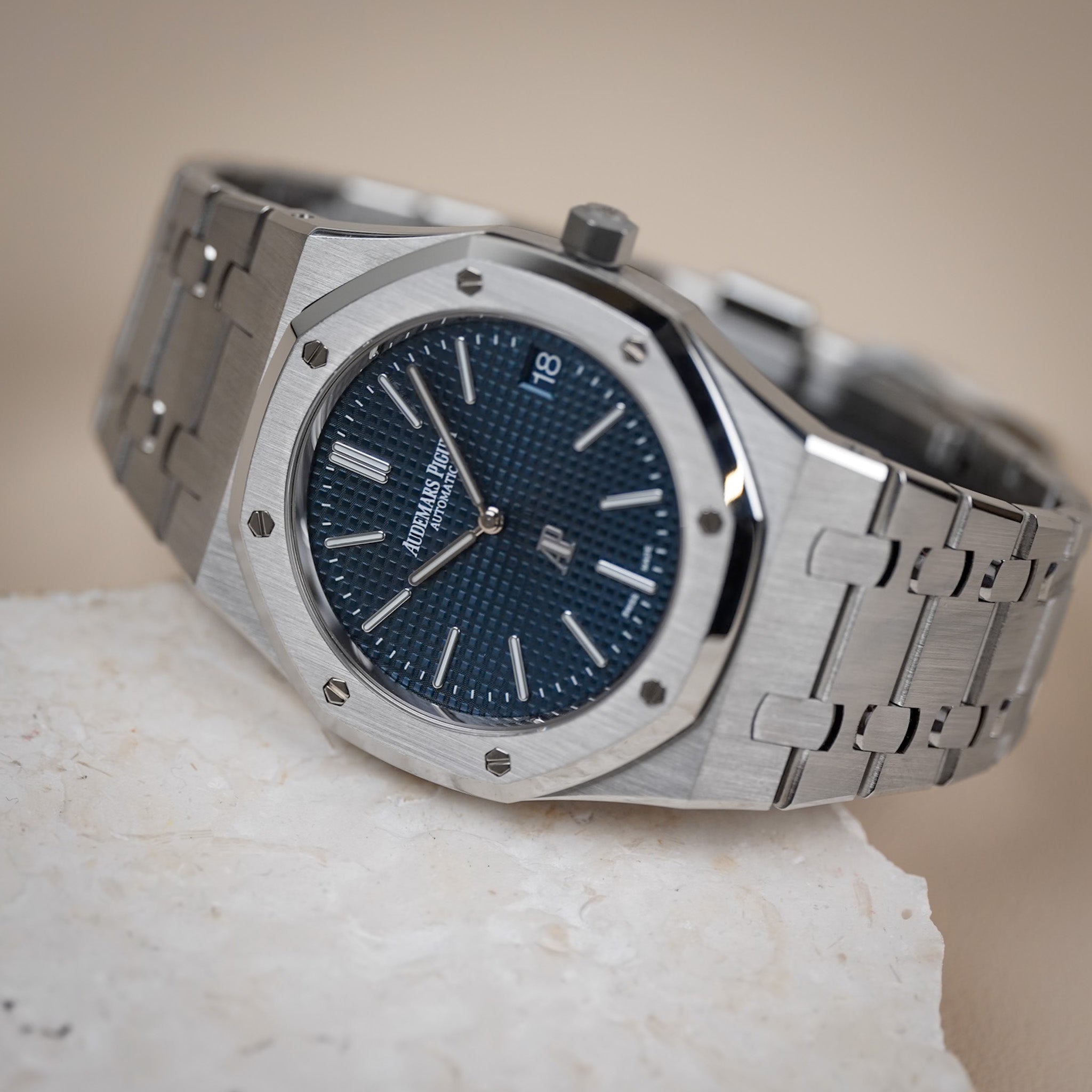 A steel Audemars Piguet Royal Oak with a textured blue dial, octagonal bezel with exposed screws, and a date window showing "18", shot lying sideways on a light stone surface