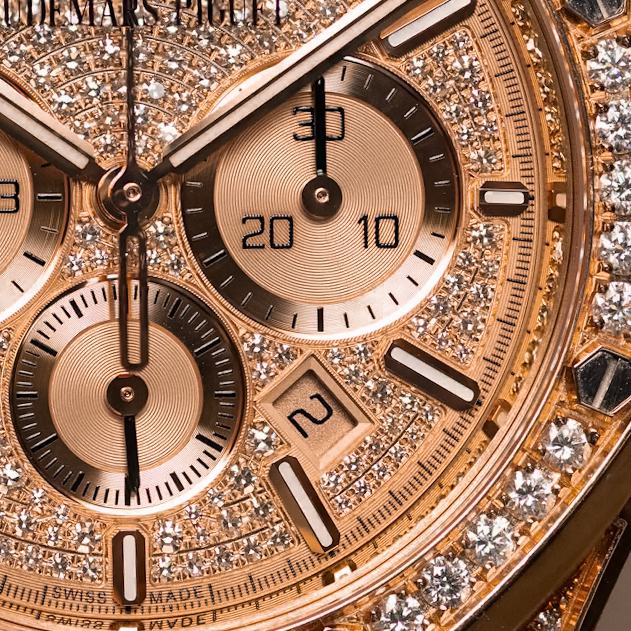 Close-up of a luxury chronograph watch dial covered in sparkling pavé stones, with rose-gold-toned subdials, baton indices, and a date window. The brand name 'AUDEMARS PIGUET' is partially visible at the top.
