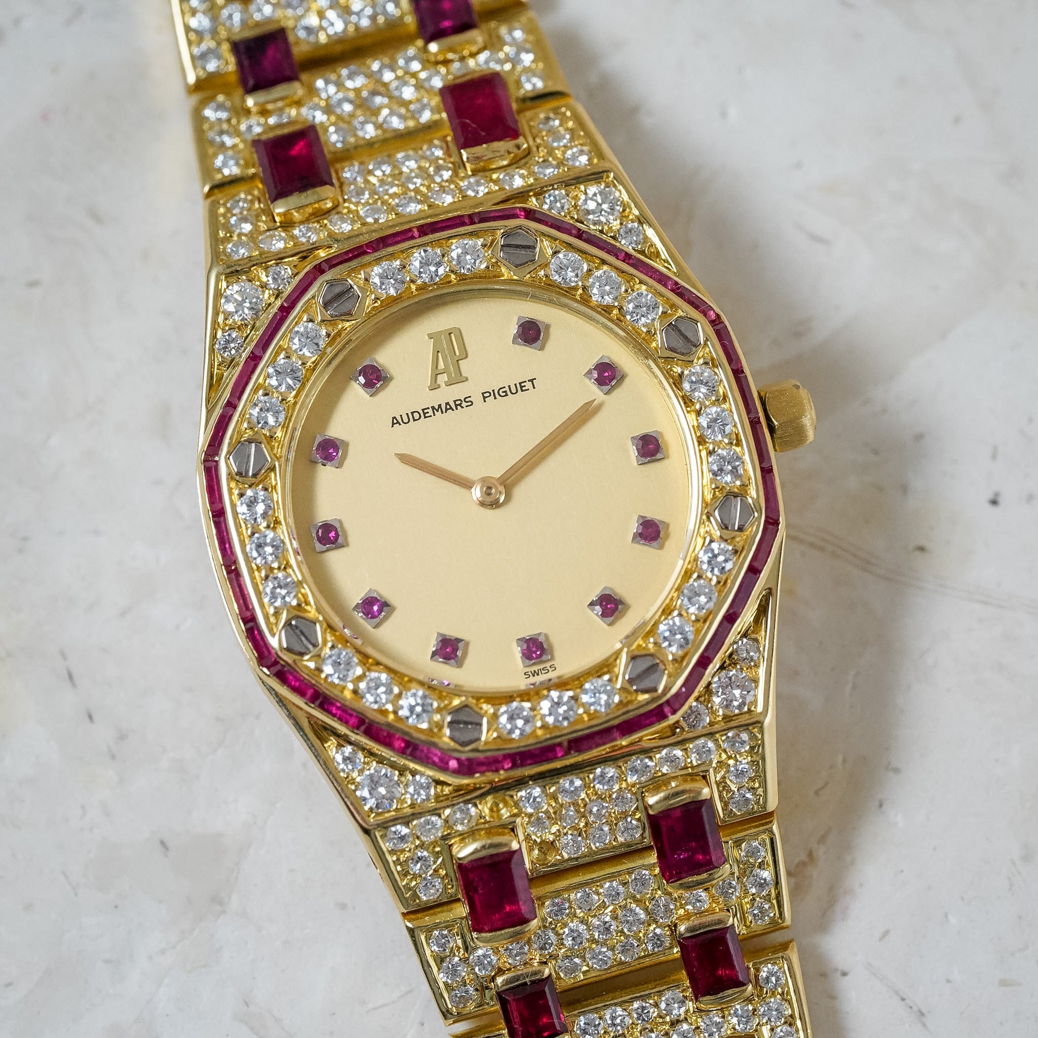 A luxury gold-plated Audemars Piguet watch featuring a diamond-encrusted bezel, a cream dial with gold-tone hands, and a link bracelet embellished with deep red ruby stones and pavé diamonds, set against a light marble background.