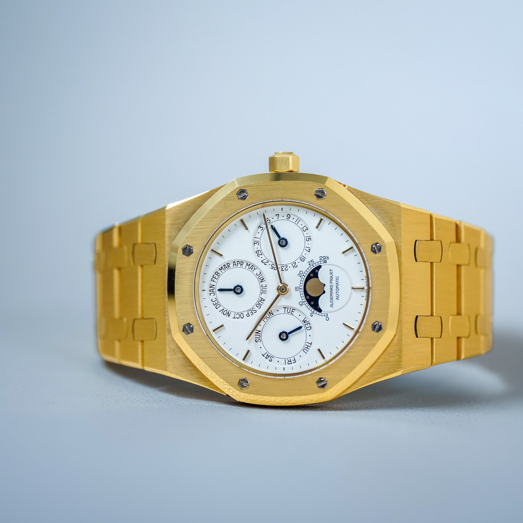 A gold wristwatch with an octagonal bezel, white dial, and matching gold link bracelet, photographed on a light blue background.