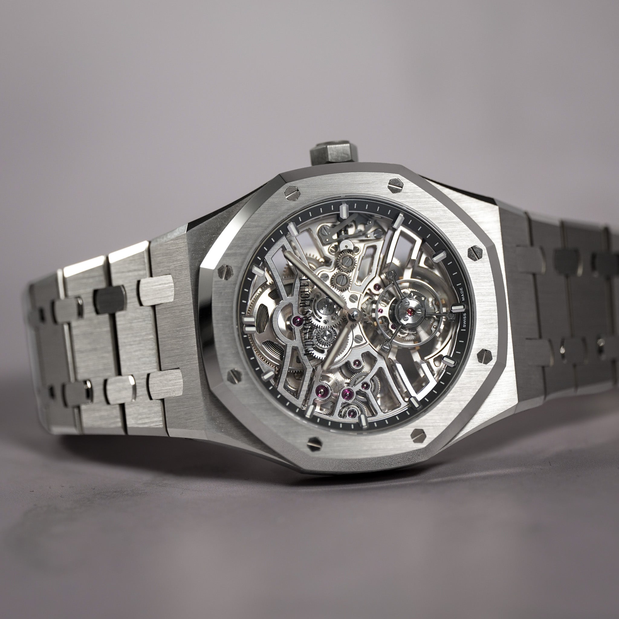 A luxury stainless steel skeleton watch with an octagonal bezel and a matching metal link bracelet, displayed on a neutral gray background.