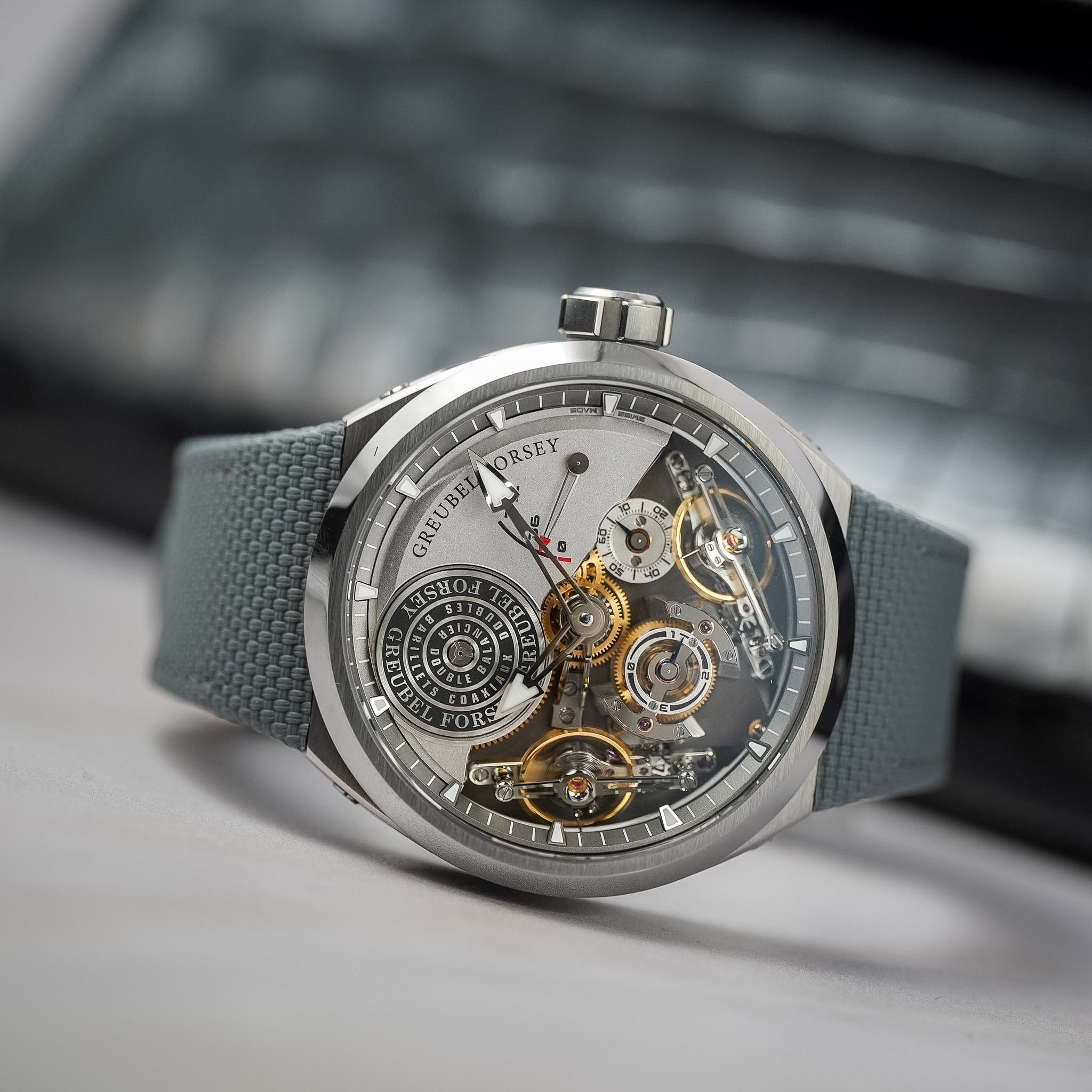 A luxury skeleton watch by featuring a titanium case, a detailed open dial showing gold and silver mechanical gears, and a textured gray strap, set against a blurred dark background.