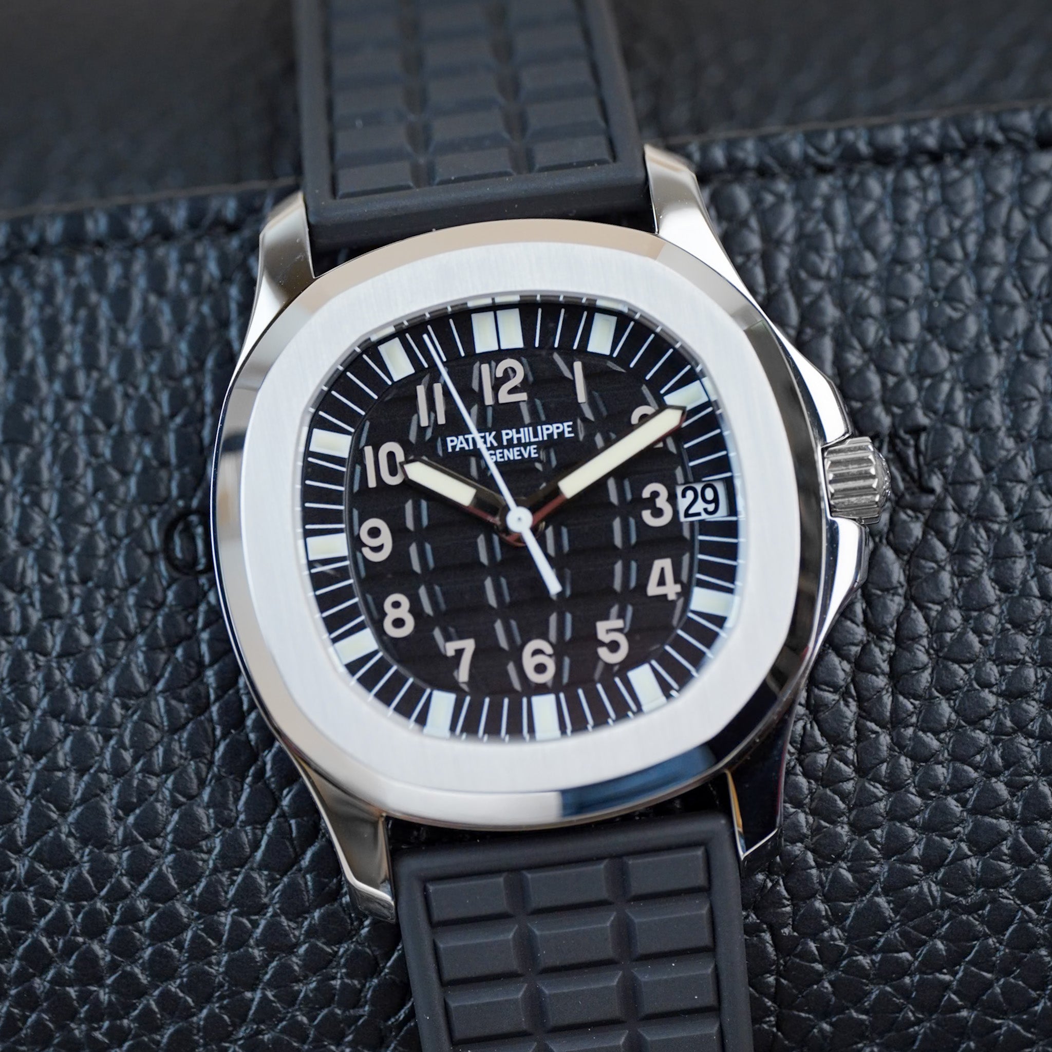 A stainless steel Patek Philippe Aquanaut sport watch with a textured black dial, textured black rubber strap, and white hands and hour markers, shot flat against a black leather surface
