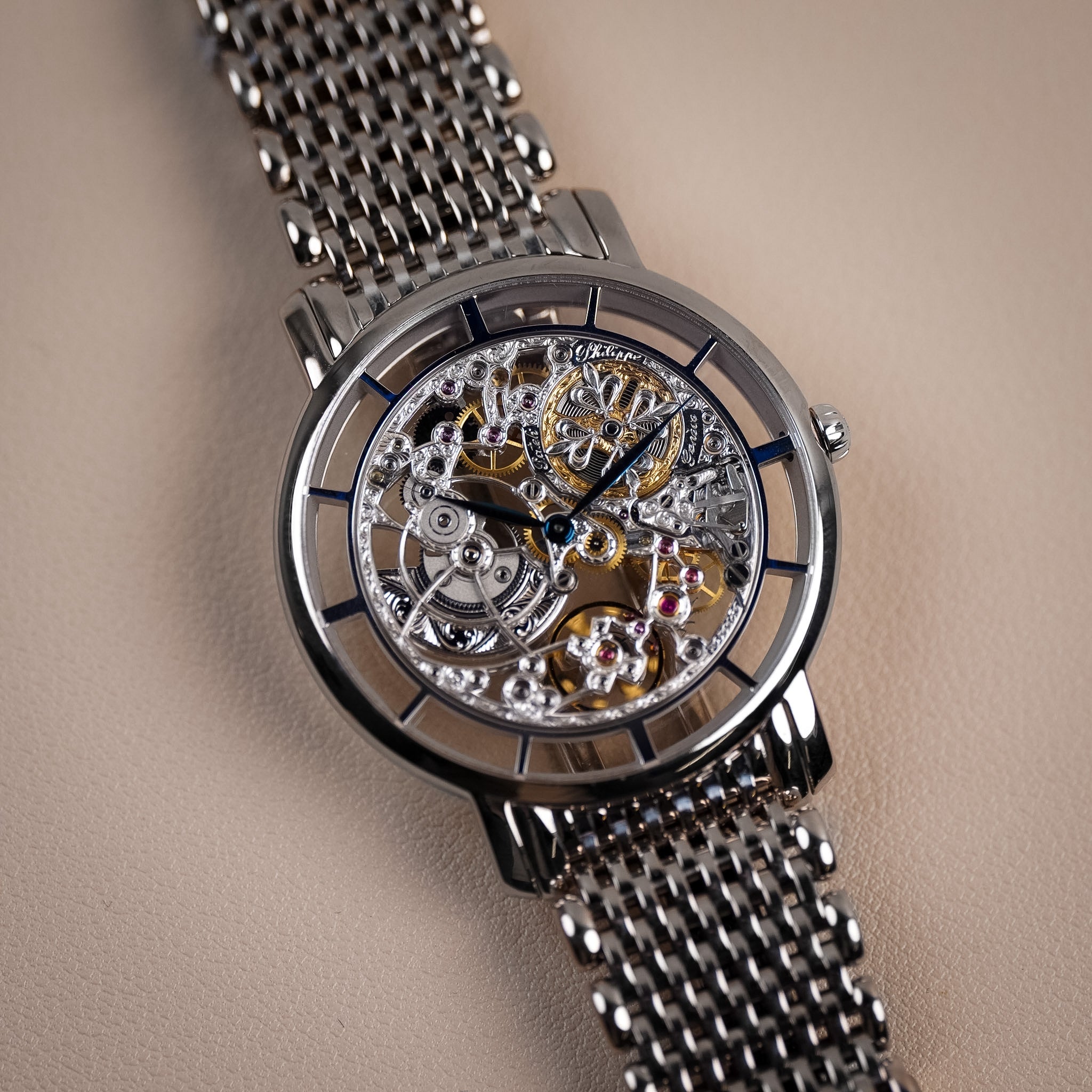 A white gold Patek Philippe watch with an intricately decorated skeletonized movement on a white gold mesh bracelet, shot against a neutral cream background