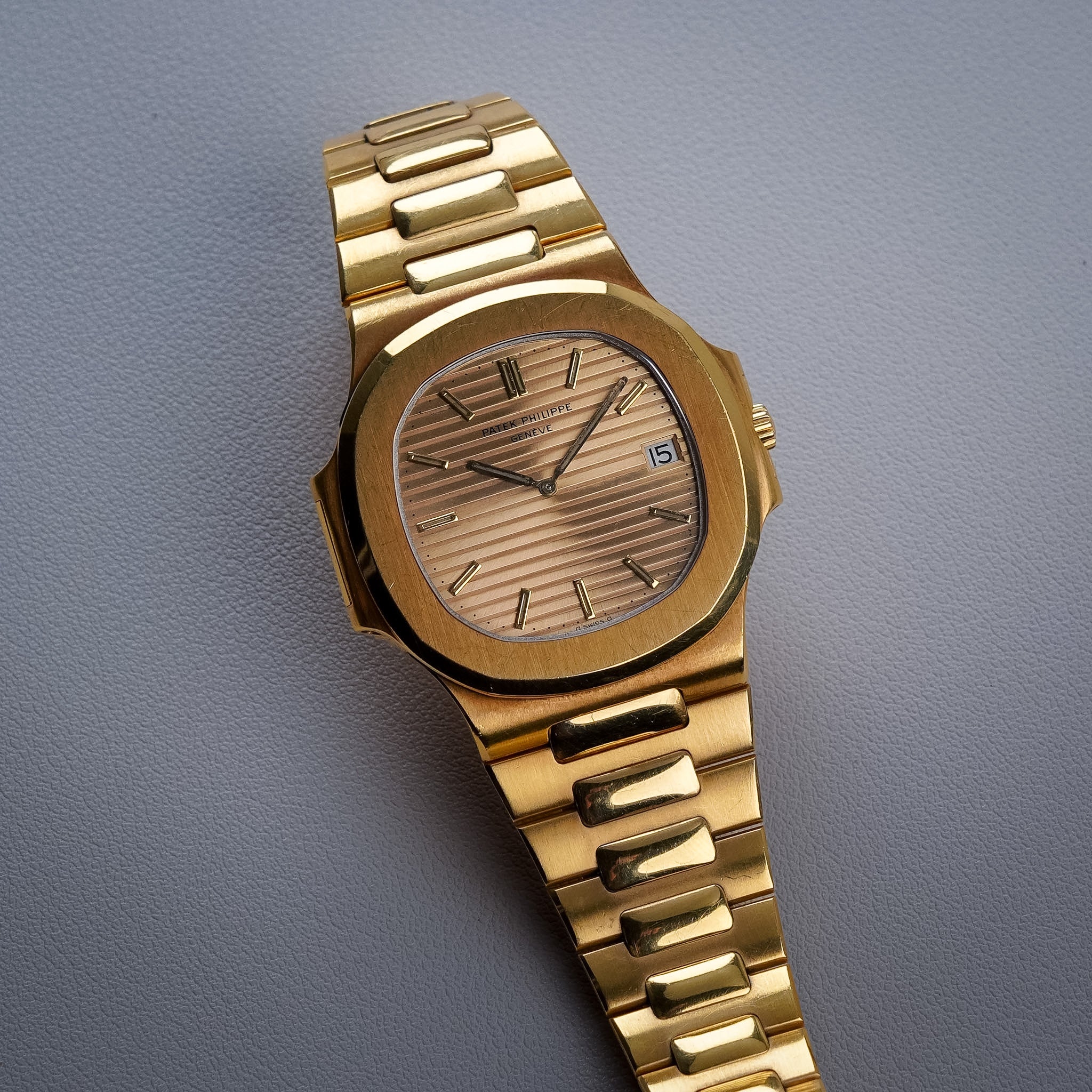 A yellow gold Patek Philippe Nautilus with a gold-tone dial, yellow gold case and bezel, gold hands and hour markers, and a date window at 3 o'clock. Shot resting on a light grey textured surface.