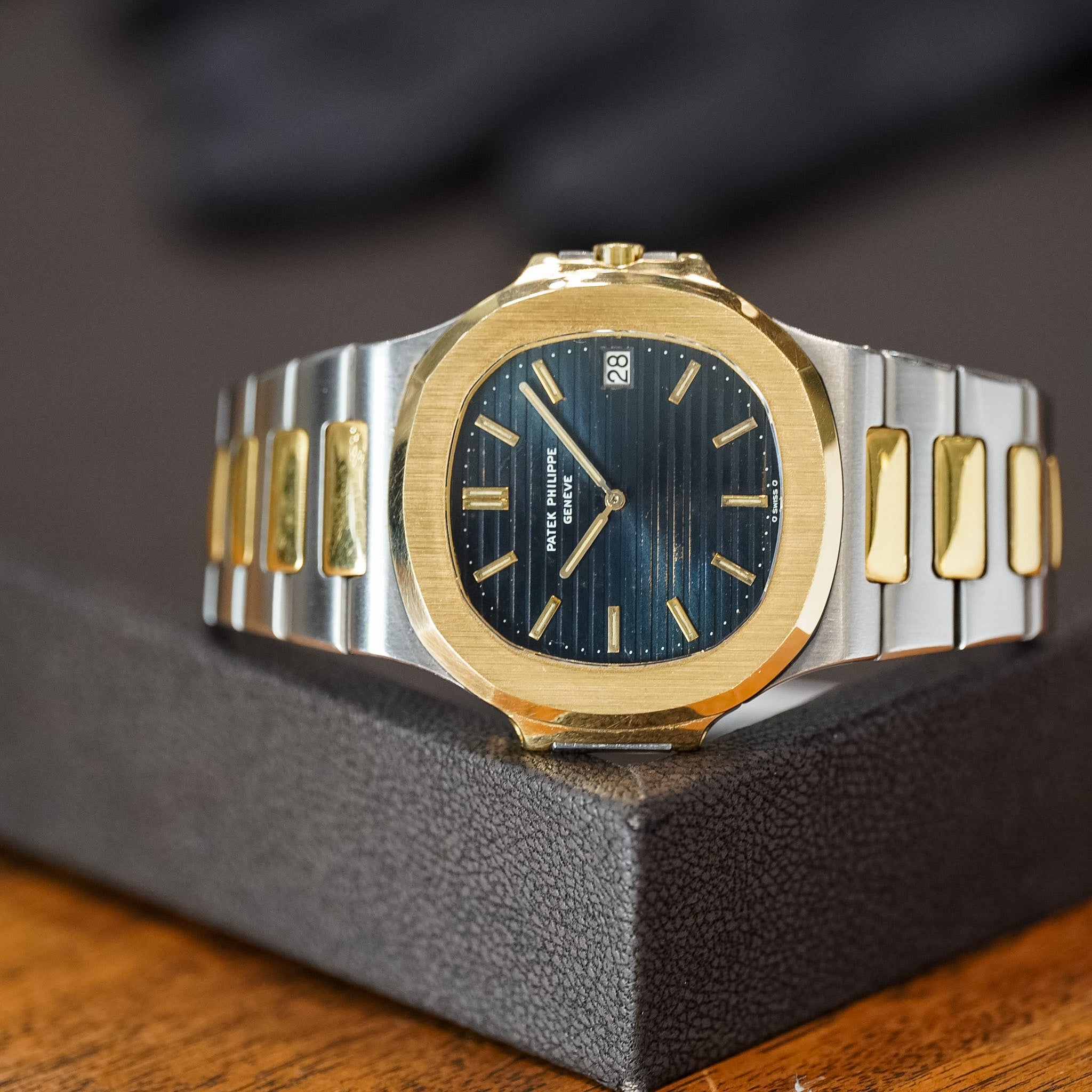 A two-tone gold and silver stainless steel watch with a dark blue sunburst dial, resting on a textured dark gray display block over a wooden surface.