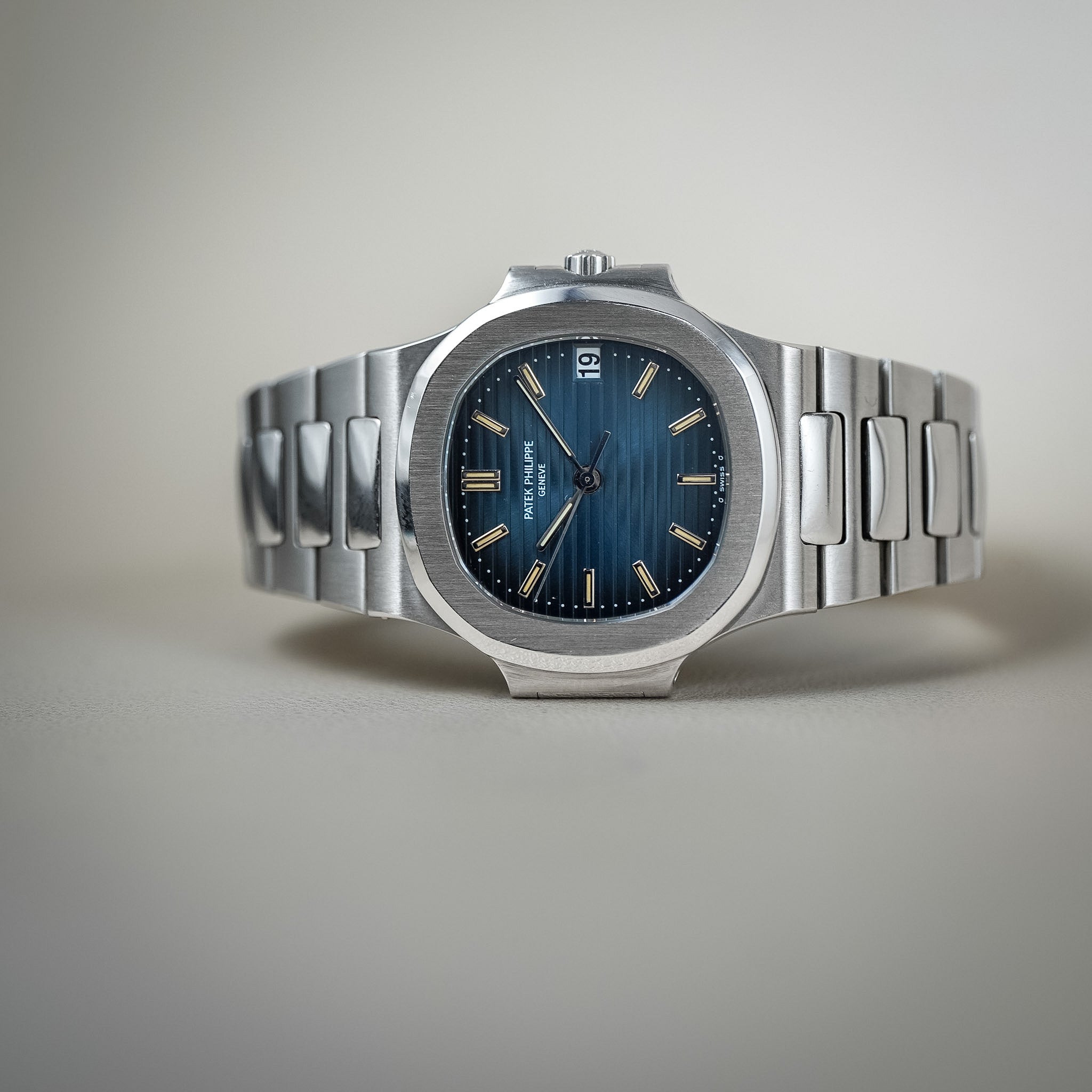 A stainless steel wristwatch with a blue sunburst dial, white gold hour markers and hands, and a date window at the 3 o'clock position. The watch is photographed on a neutral light gray background.