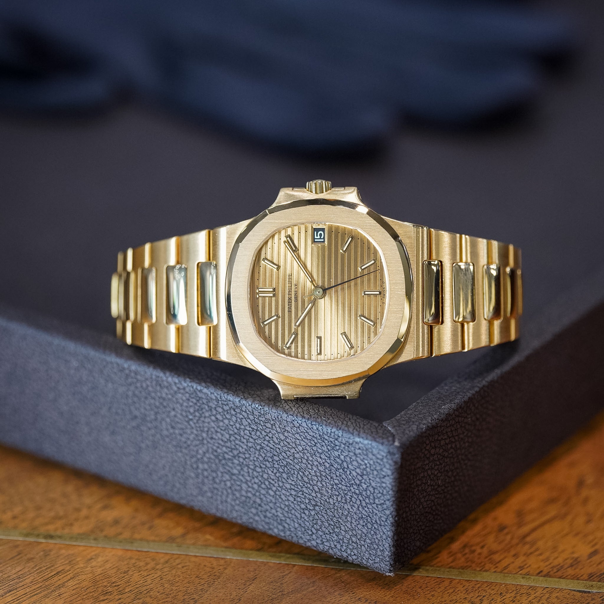 A gold wristwatch with an octagonal bezel, matching gold dial, and integrated metal bracelet, displayed on a dark textured tray over a wooden surface.