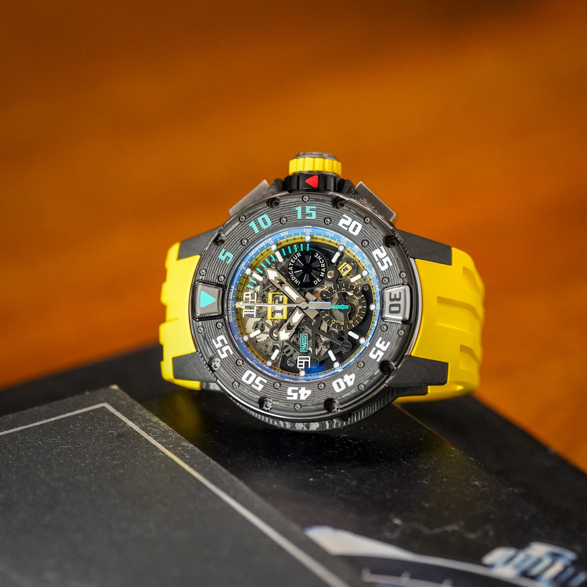 A luxury chronograph watch with a black and yellow color-blocked case, a yellow rubber strap, and a highly detailed skeleton dial, resting on a dark surface with a warm orange background.