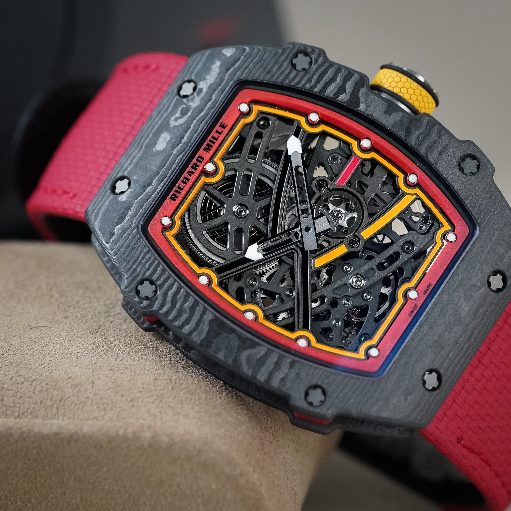 A close-up of a Richard Mille wristwatch with a textured dark gray carbon fiber case, a skeletonized dial showing black gears, and a red and yellow inner ring. The watch is worn on a wrist with a bright red textured strap.