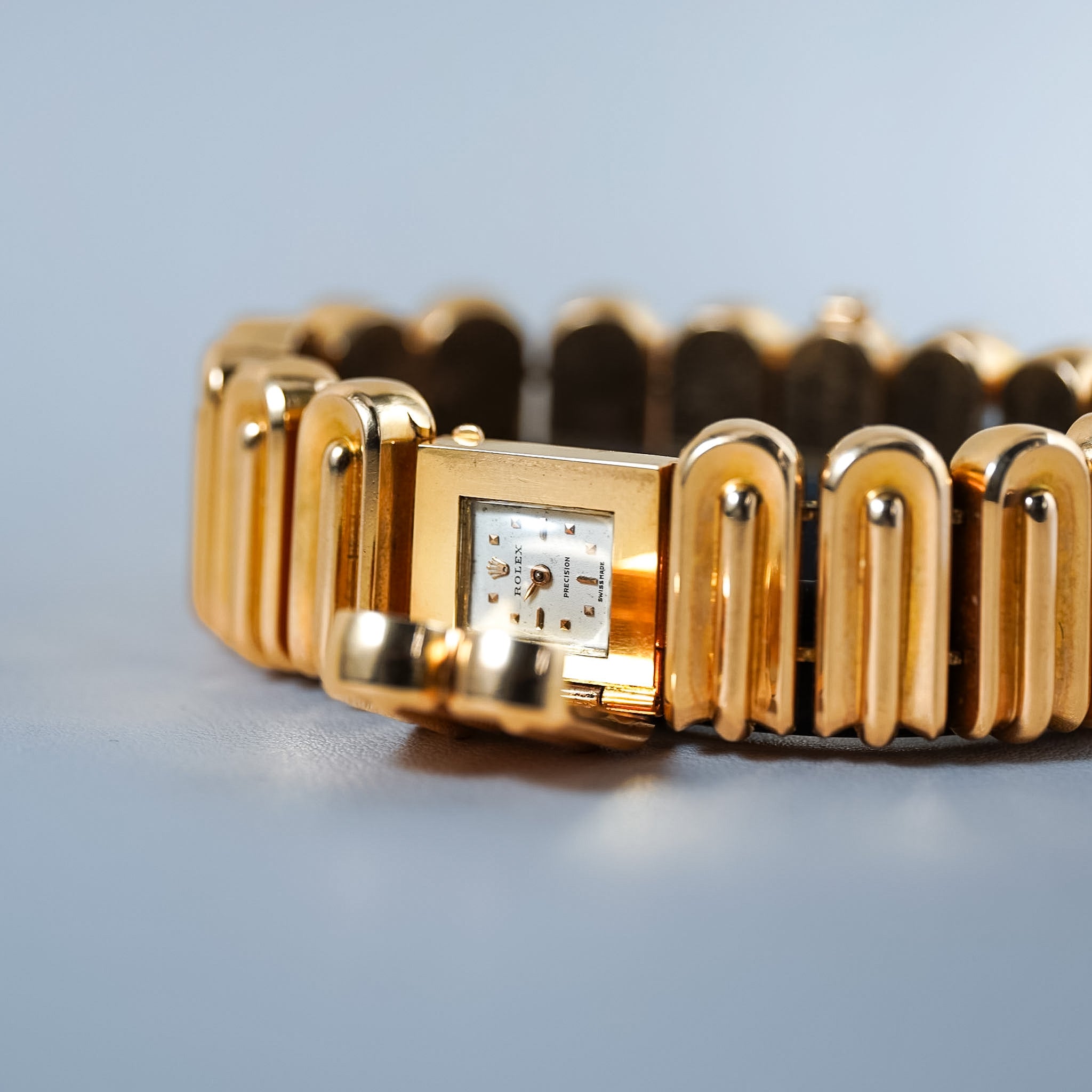 A gold-tone rectangular watch with a segmented link bracelet, shown in a close-up studio shot against a soft blue-gray background.