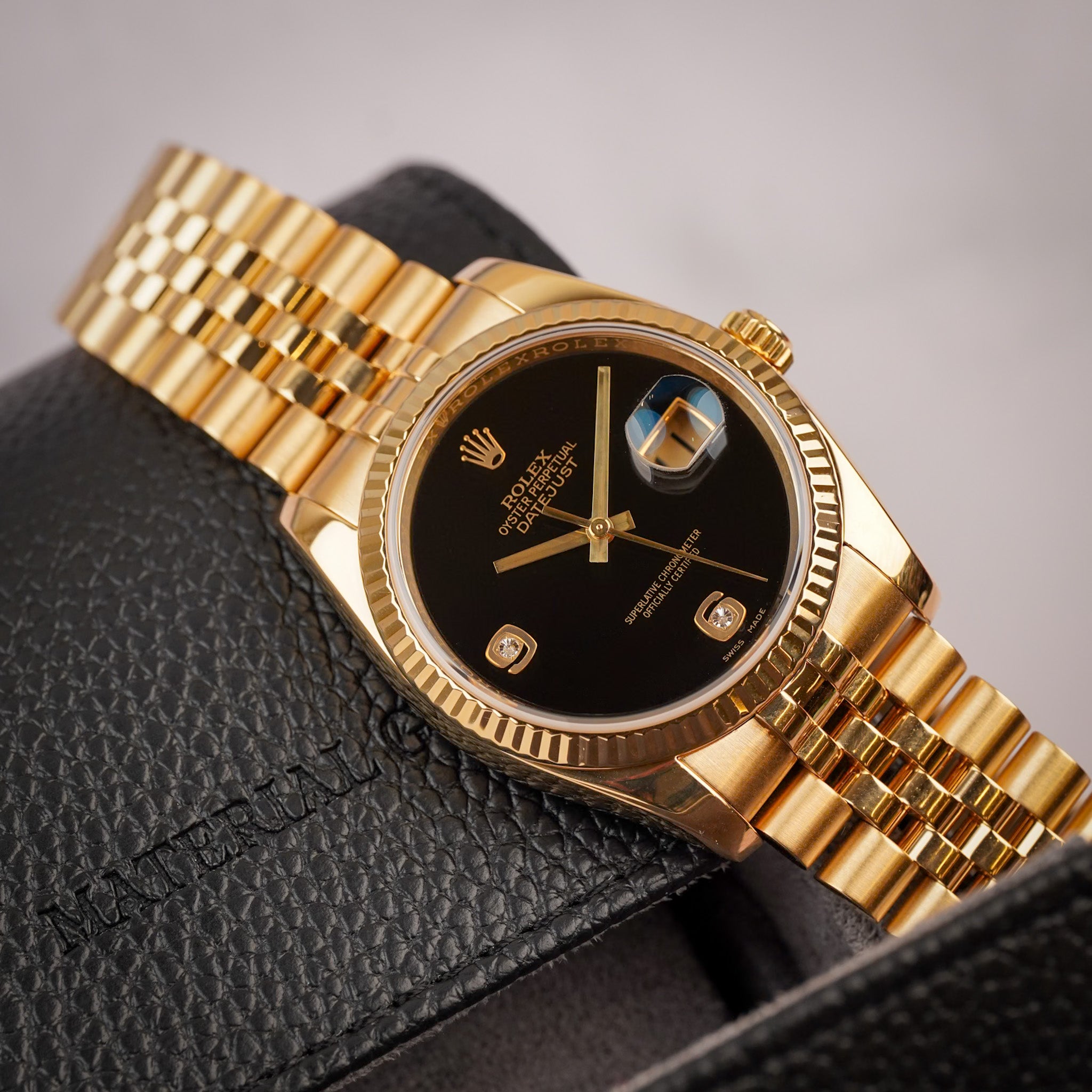 A yellow gold Rolex Datejust with a fluted bezel, deep black onyx dial, gold Jubilee bracelet, and a date window showing "11", resting diagonally on a black leather case.