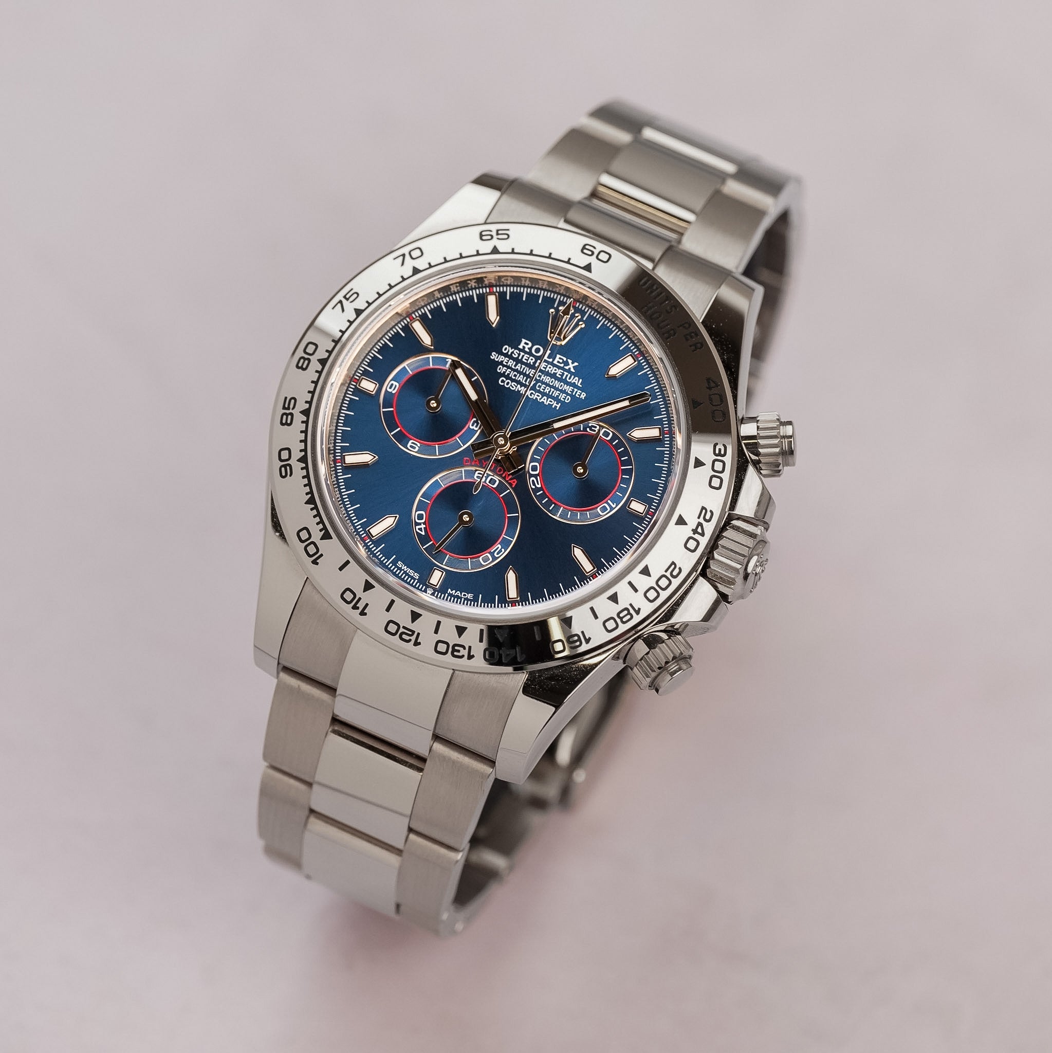 A white gold Rolex Daytona with a blue dial, red accents, and a white gold bracelet, shot at an angle on a neutral grey table