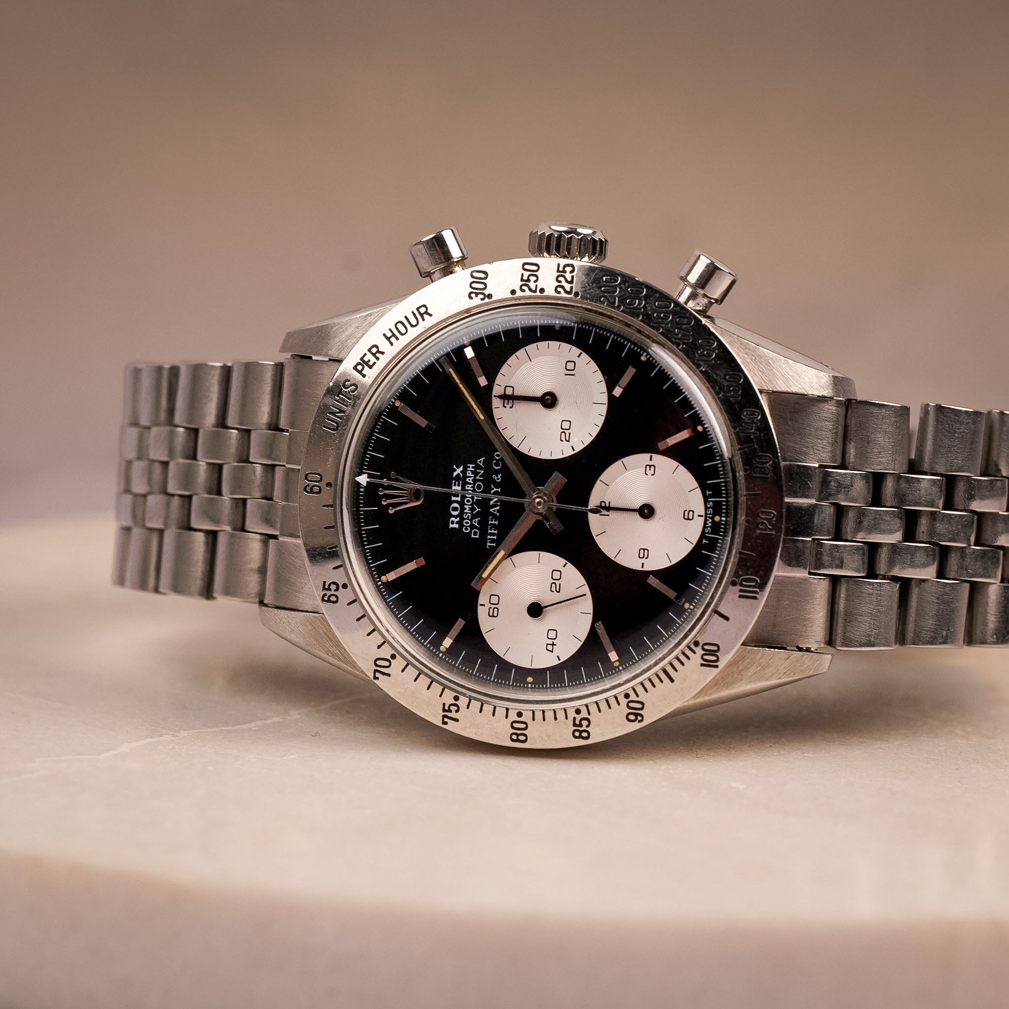 A steel vintage Rolex Daytona with a lack dial, white subdials, steel bezel, and steel bracelet, shot resting sideways on a beige stone surface with a beige background.