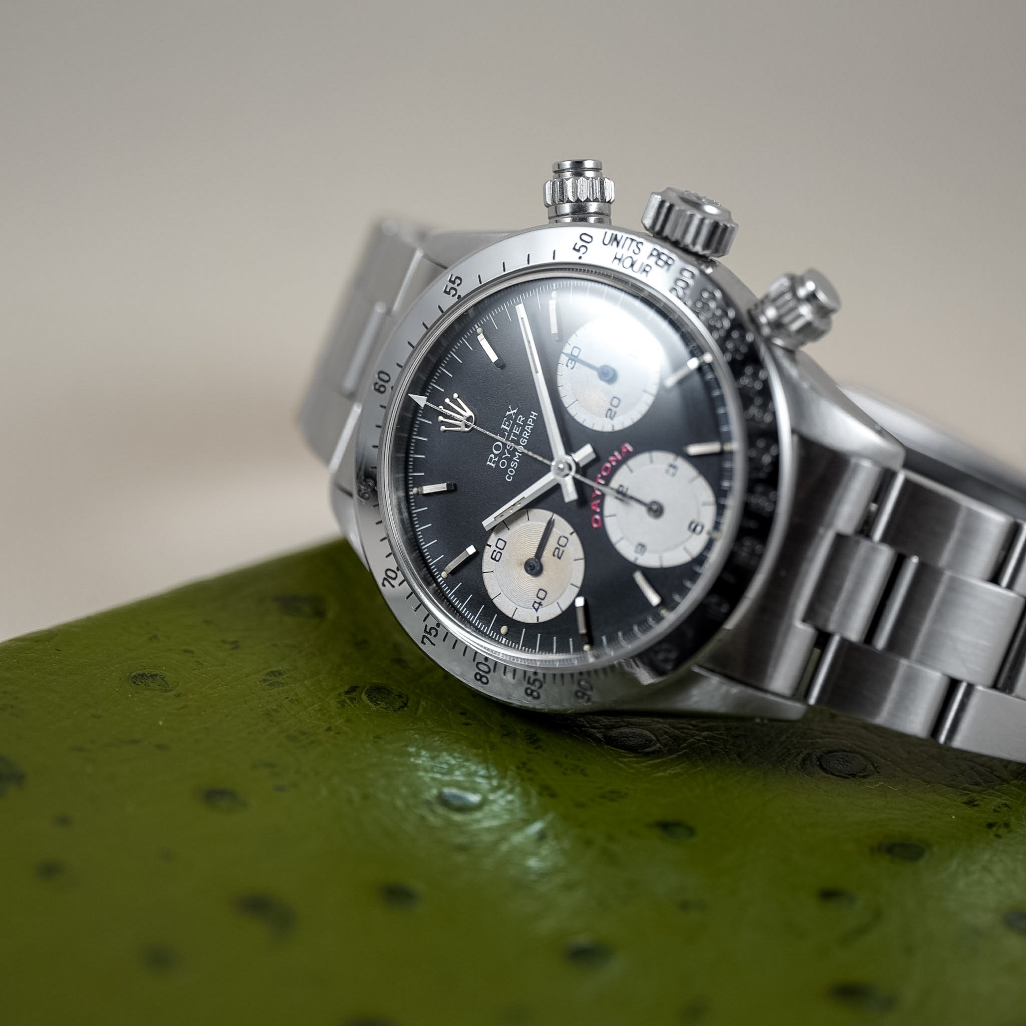 A Rolex Daytona wristwatch with a silver stainless steel Oyster bracelet and a black dial with cream subdials, resting on a textured green surface against a neutral beige background.