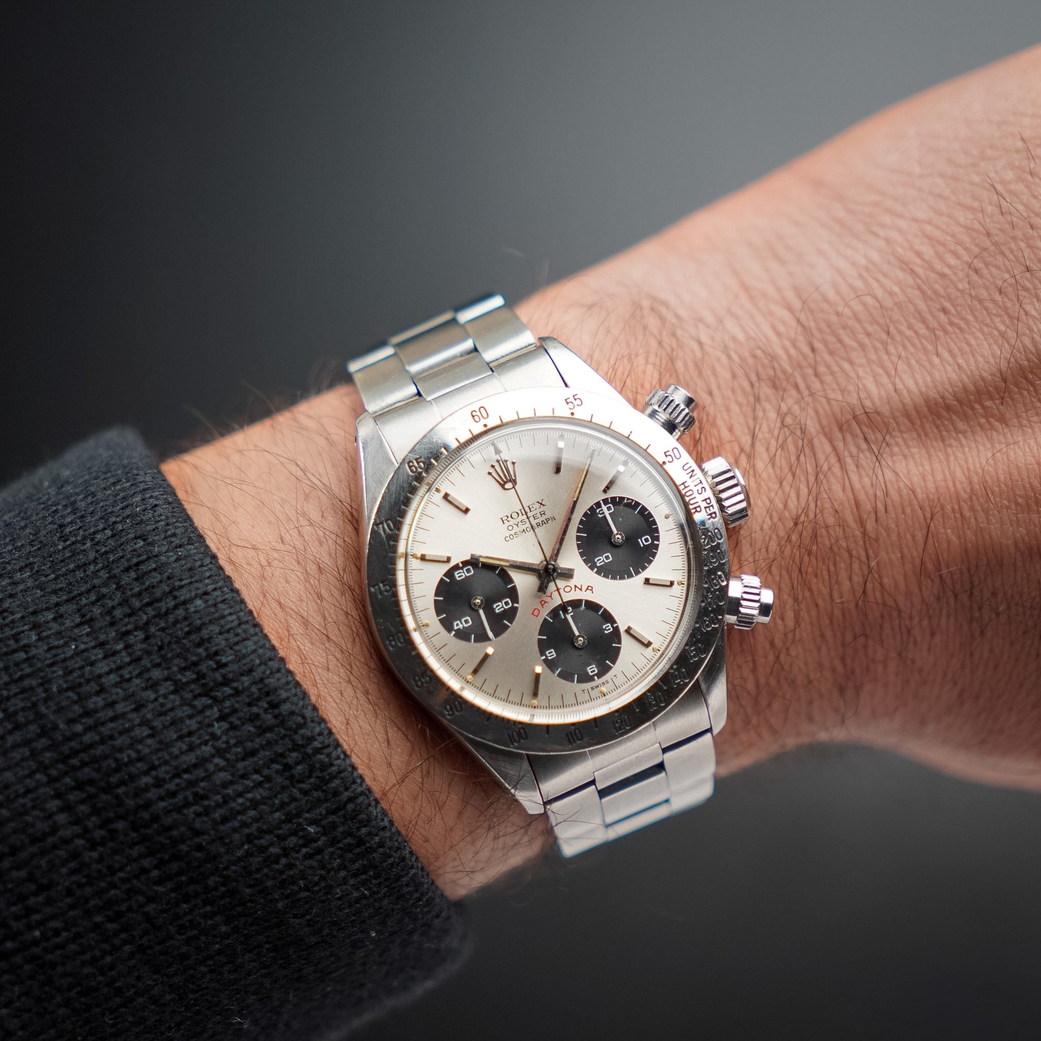A man's wrist with a steel vintage Rolex chronograph featuring a steel bezel and steel bracelet. The wrist is floating over a grey gradient background. 