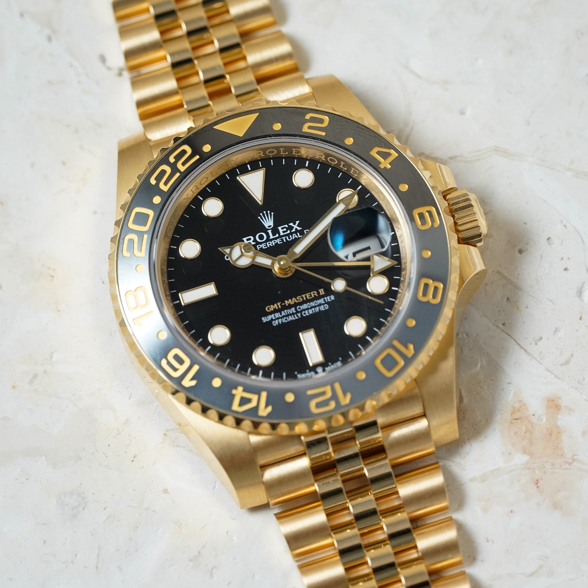 A gold Rolex GMT-Master II watch with a black dial and black bezel featuring gold numerals, resting on a light textured stone surface.