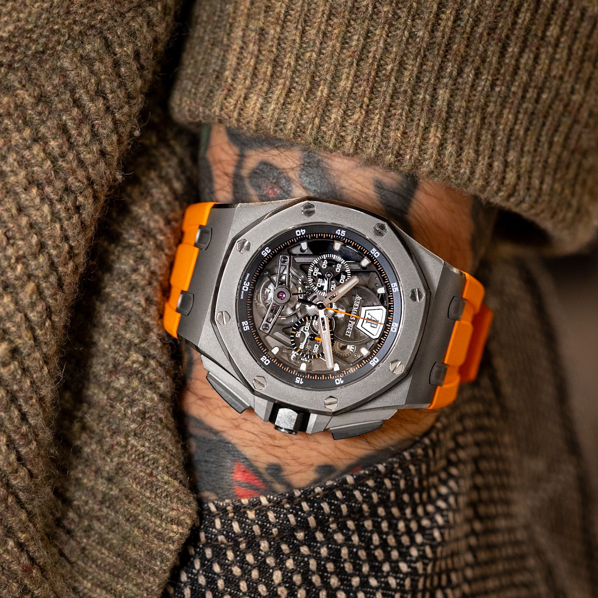 Luxury watch with orange accents on a wrist with tattoos, wearing a brown textured sweater.