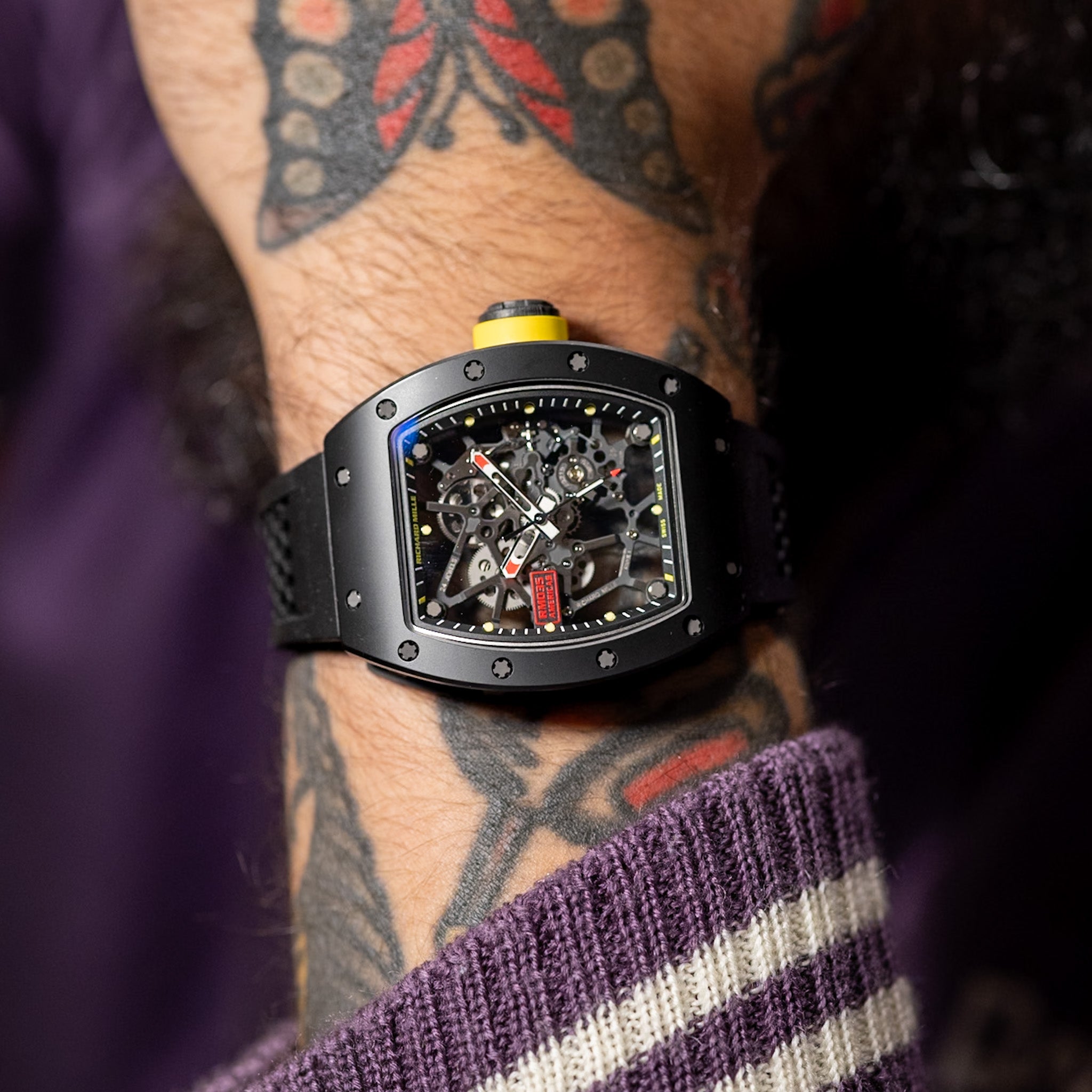 Close-up of a person's wrist wearing a black skeleton watch with visible tattoos.