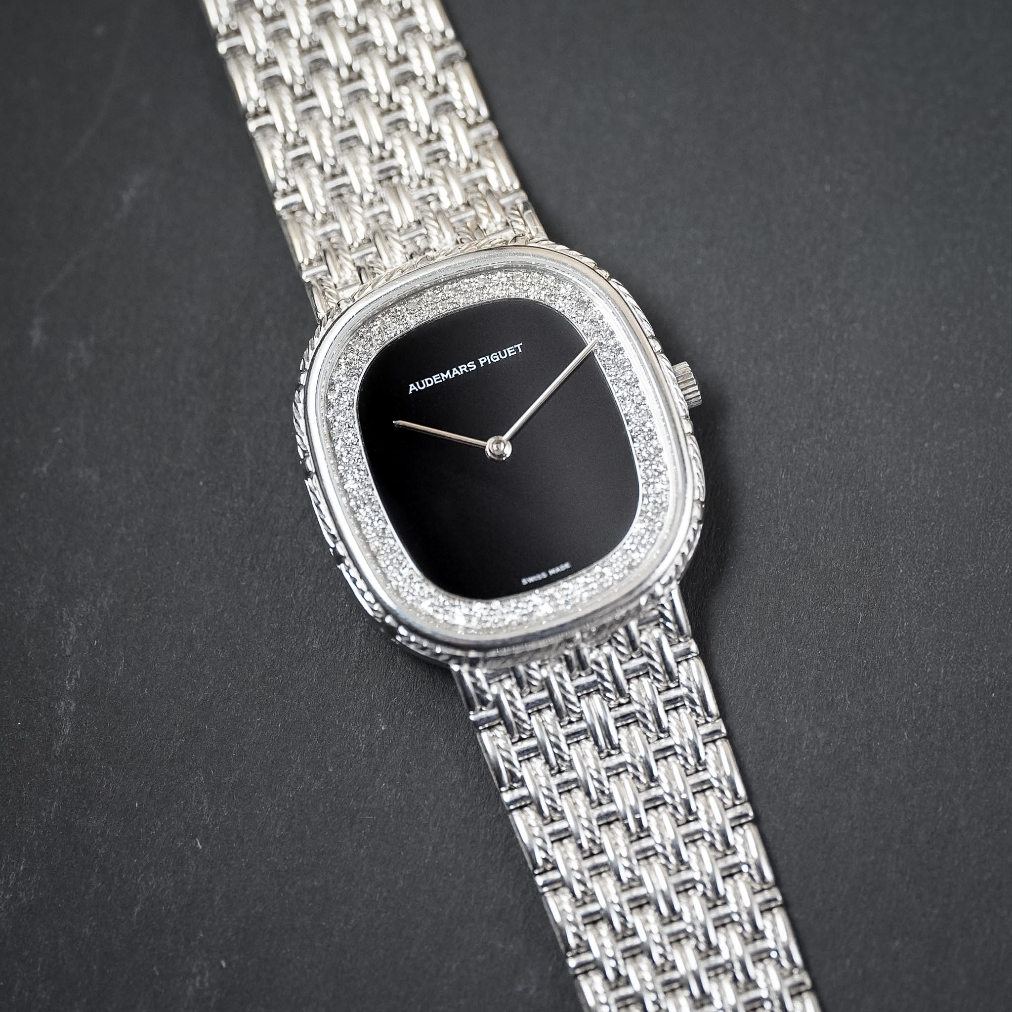 Audemars Piguet silver-tone watch with a woven bracelet, diamond-encrusted bezel, and a black dial, shown on a dark textured background.