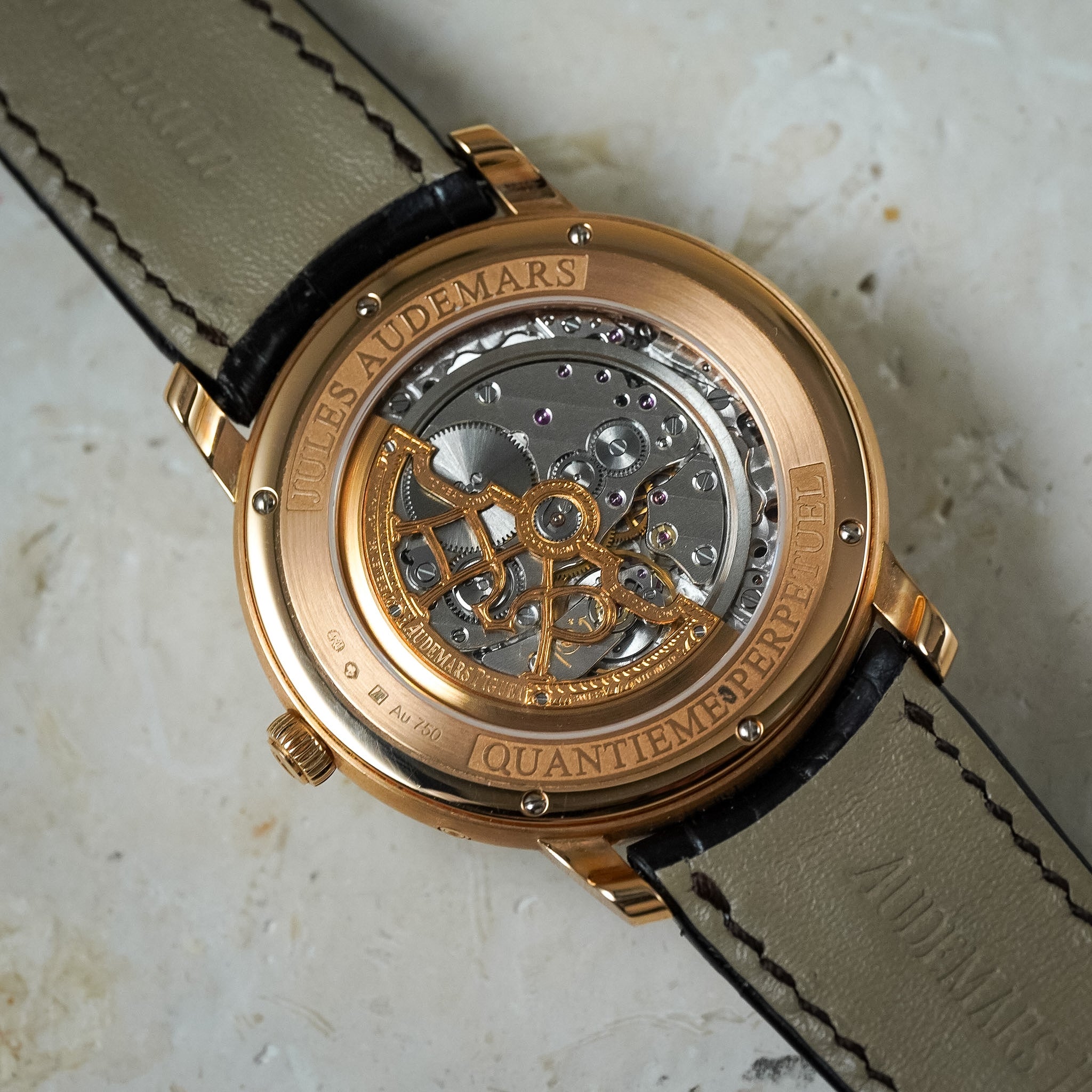 Close-up of the back of an Audemars wristwatch with a rose gold case, transparent side window showing the mechanical movement, and a taupe leather strap on a light stone surface.