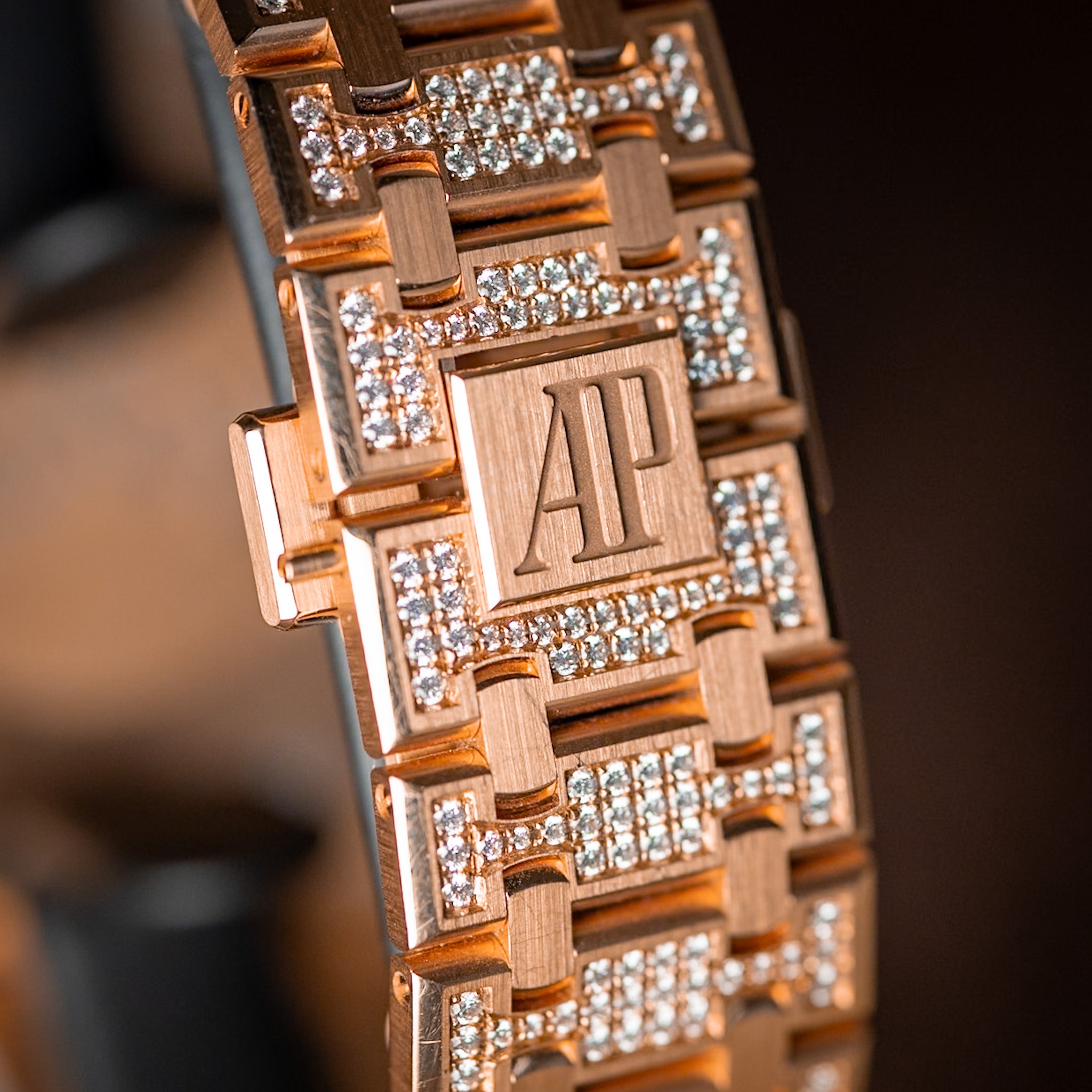 Close-up of a rose gold bracelet with pavé-set clear stones and an engraved AP logo on the center link, shown against a dark blurred background.