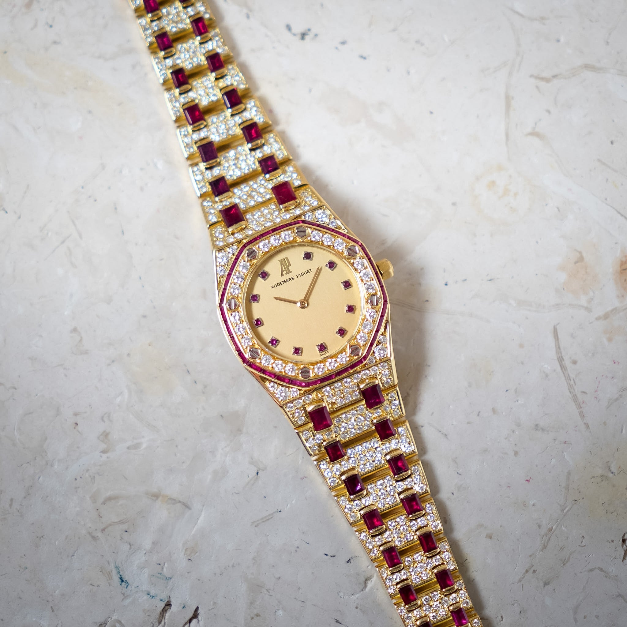 A gold-plated wristwatch with a diamond-encrusted link bracelet and red gemstone accents, photographed on a textured light beige stone surface.