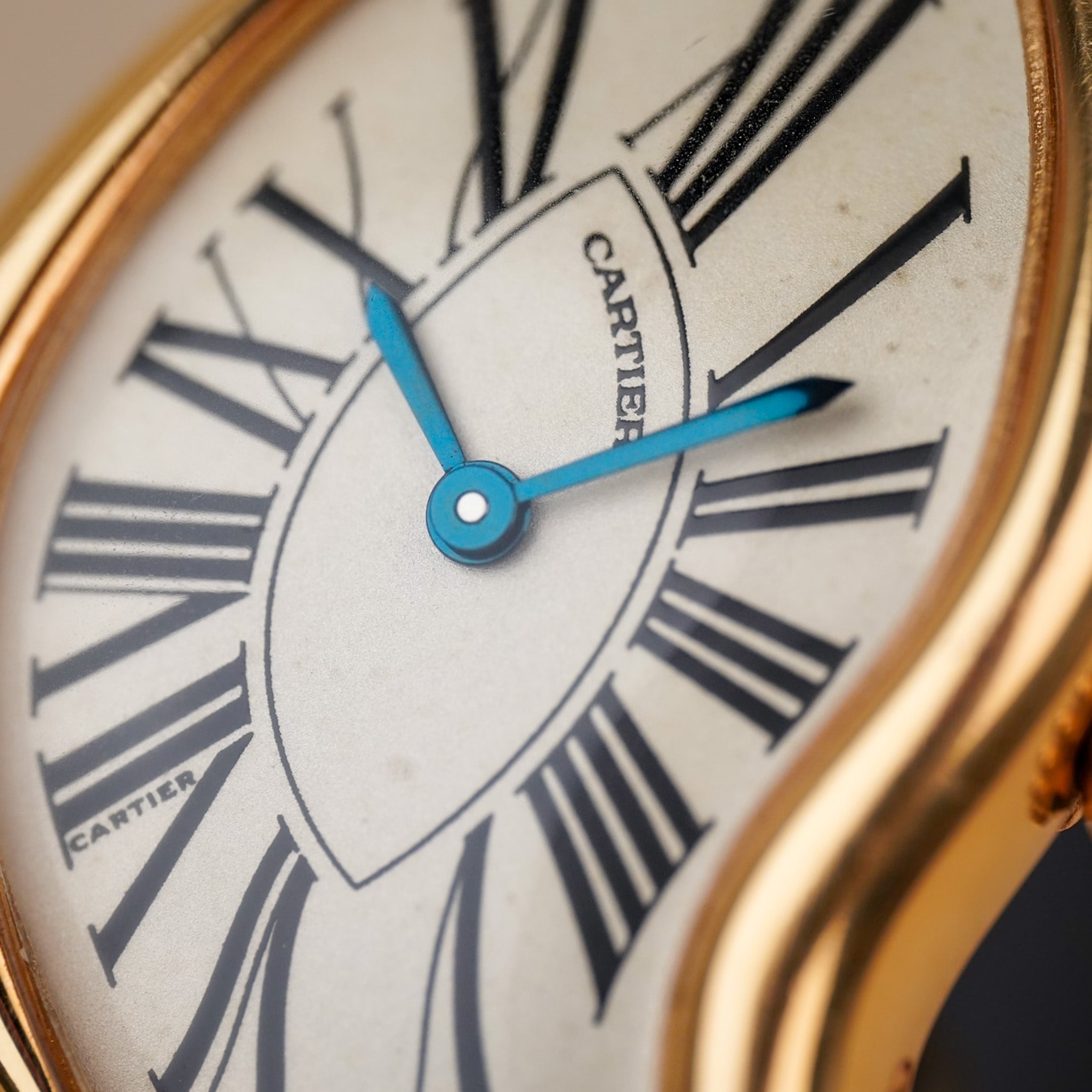 Close up of an asymmetrical Cartier watch with a white dial, black roman numeral hour markers, blue hands, a yellow gold case, and two "Cartier" logos on the dial.