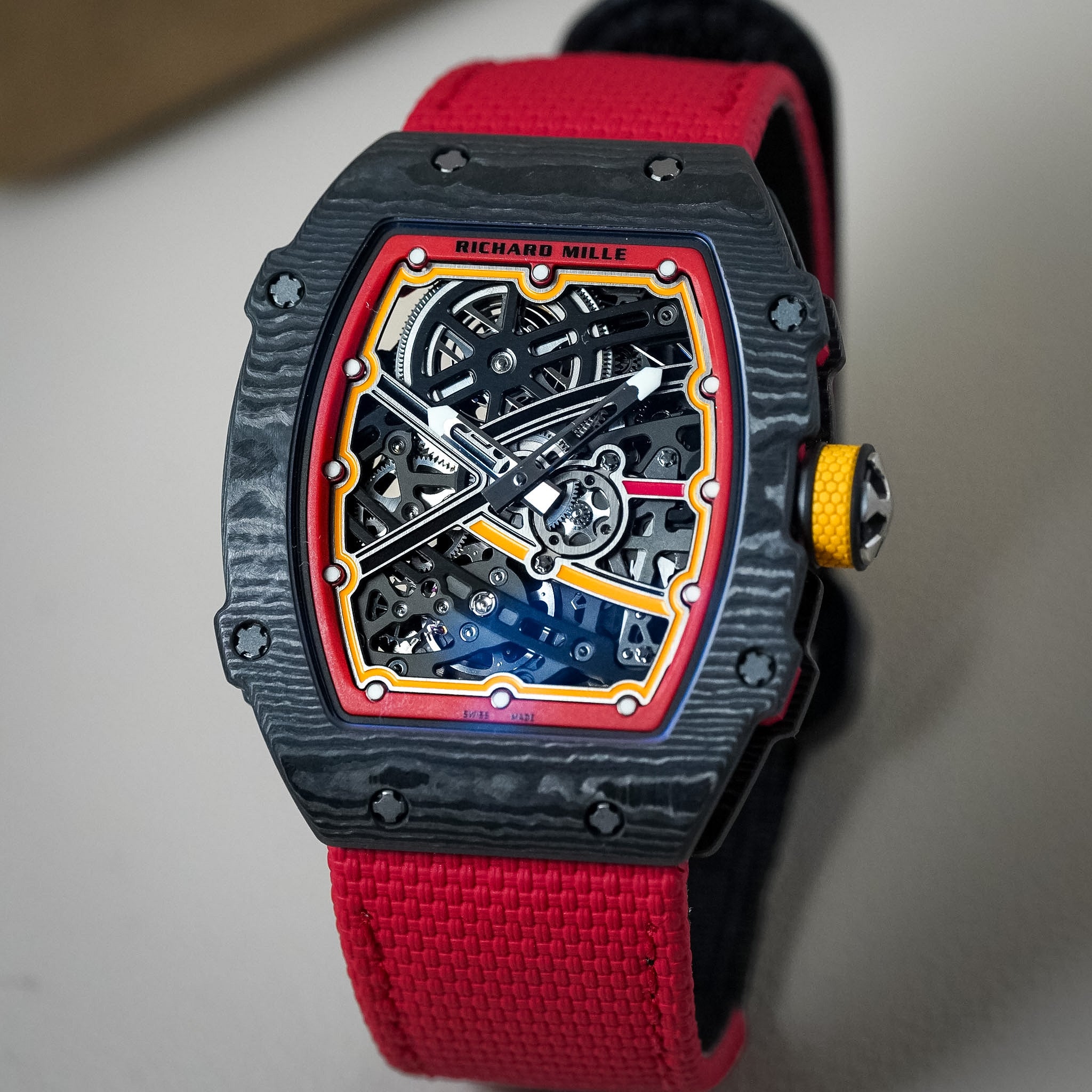 A Richard Mille wristwatch with a black textured carbon case, a skeleton dial showing intricate inner gears, and a vibrant red fabric strap.