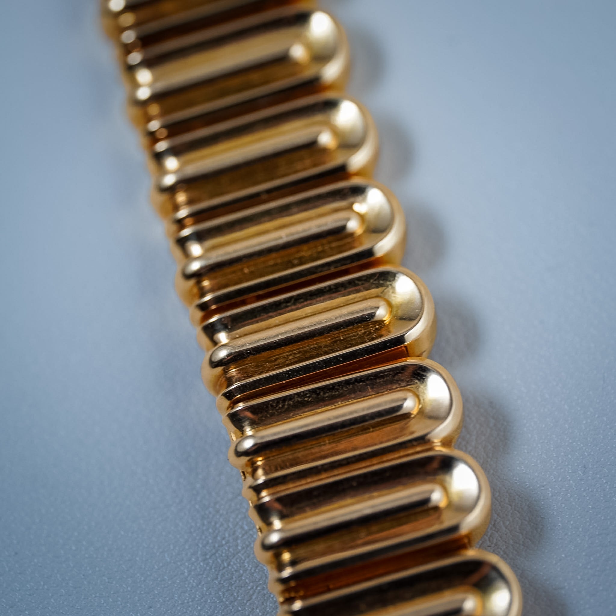 A close-up of a metallic gold chain or link accessory arranged diagonally on a smooth light blue-gray surface.