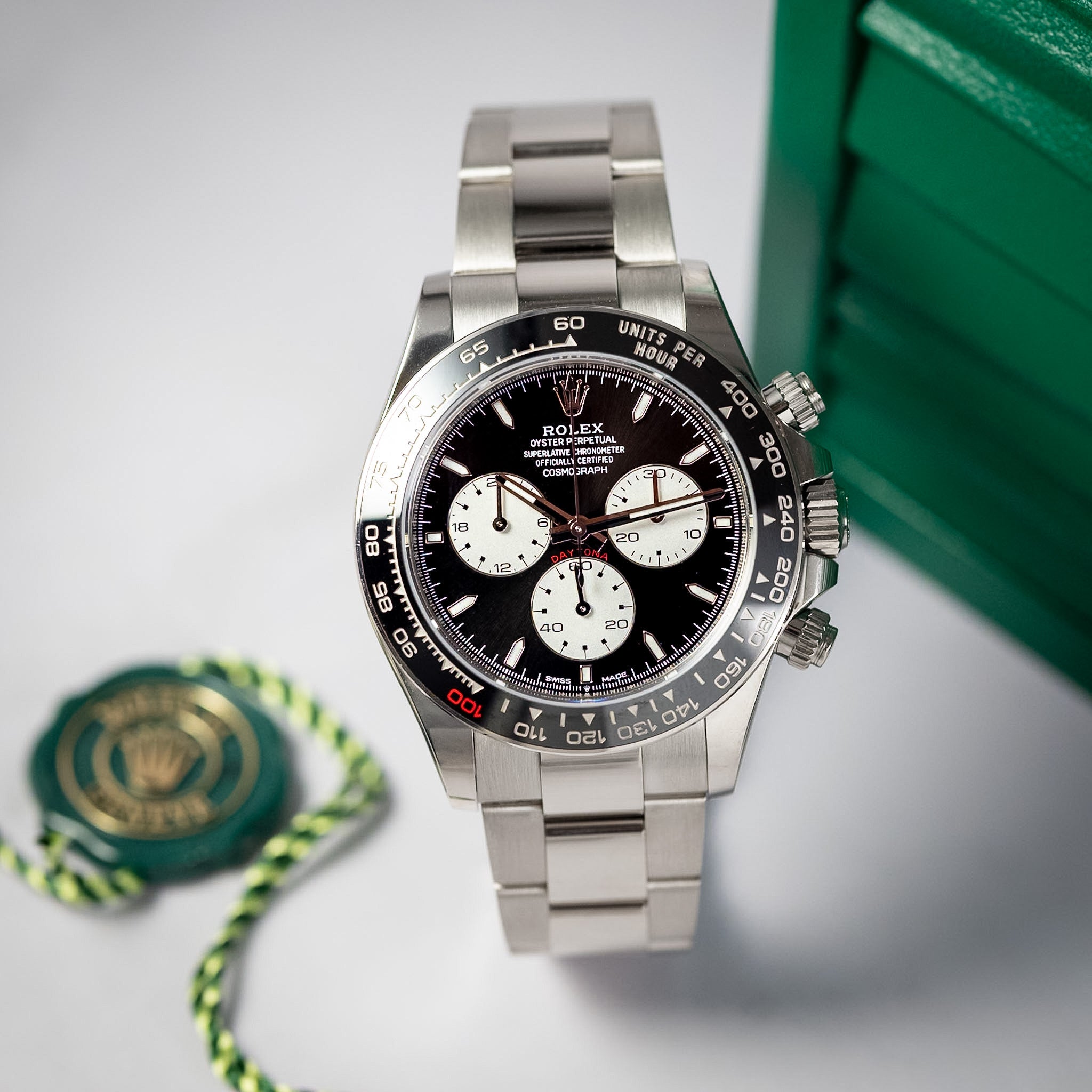A Rolex Daytona watch with a silver stainless steel bracelet, black dial, and white subdials, displayed next to its green presentation box and paper seal on a light gray background.