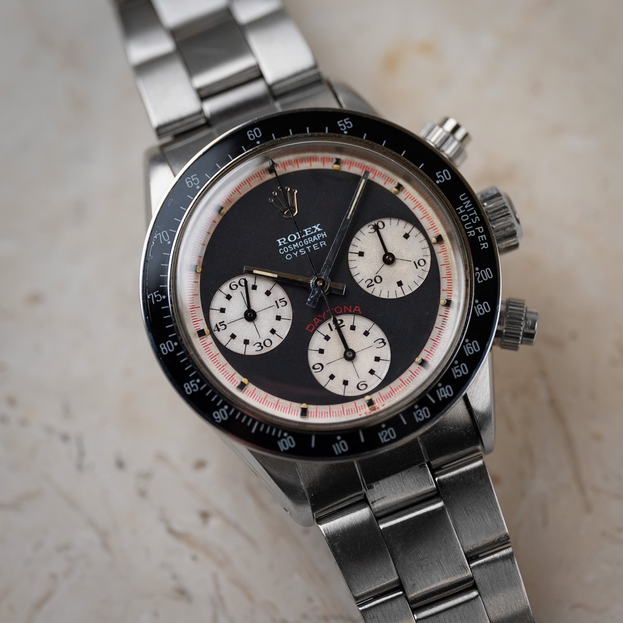 A black dial vintage Rolex Daytona in steel with a black bezel, white subdials, and red accents, shot against a cream colored stone surface.