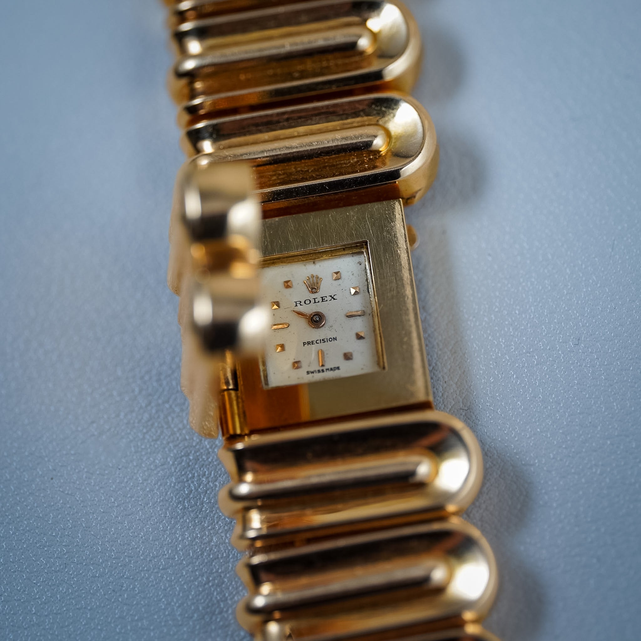 A close-up shot of a vintage Rolex Precision watch with a gold-tone metal link bracelet and a white dial, set against a textured light blue background.
