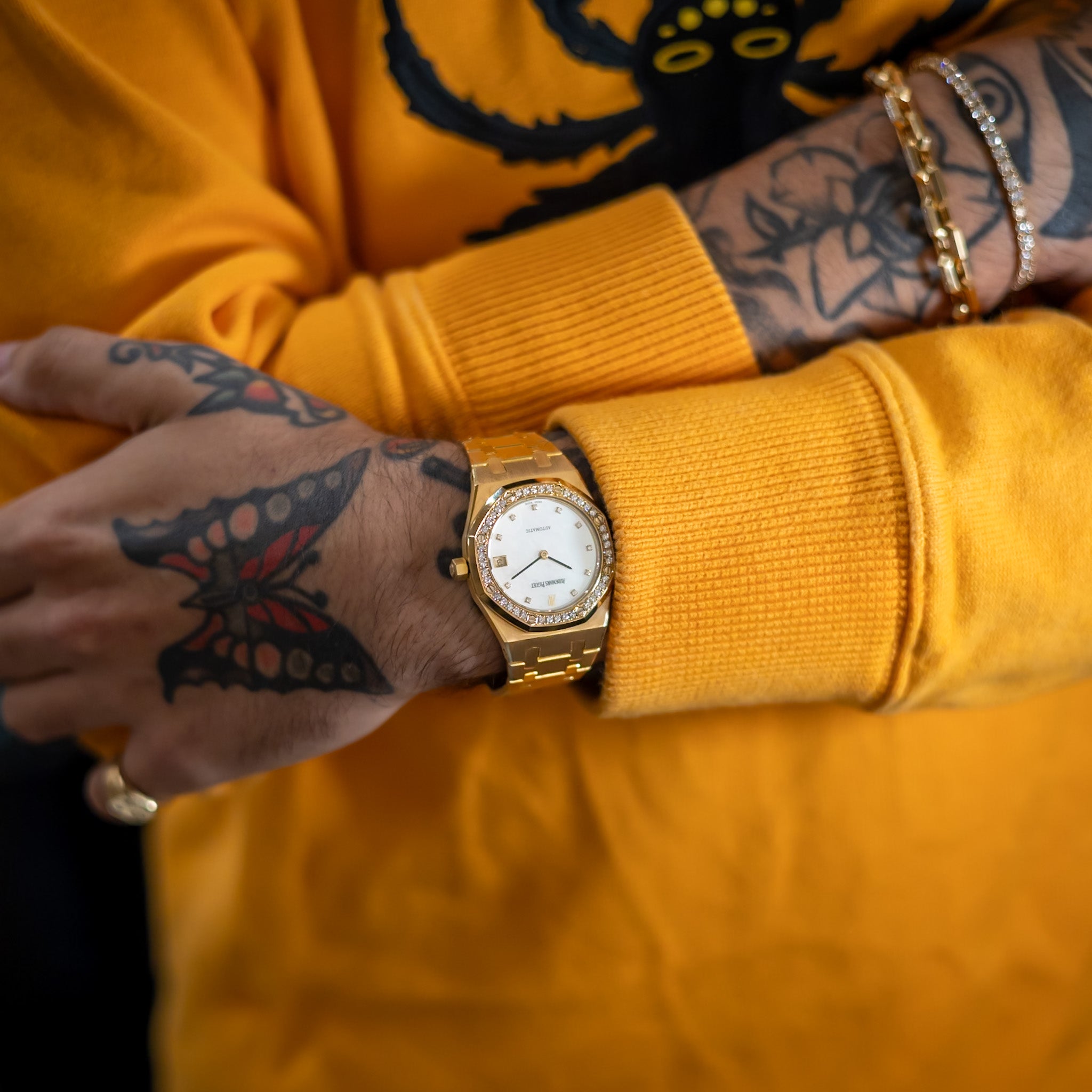 Person wearing a gold watch with a yellow sweater and tattooed arms.