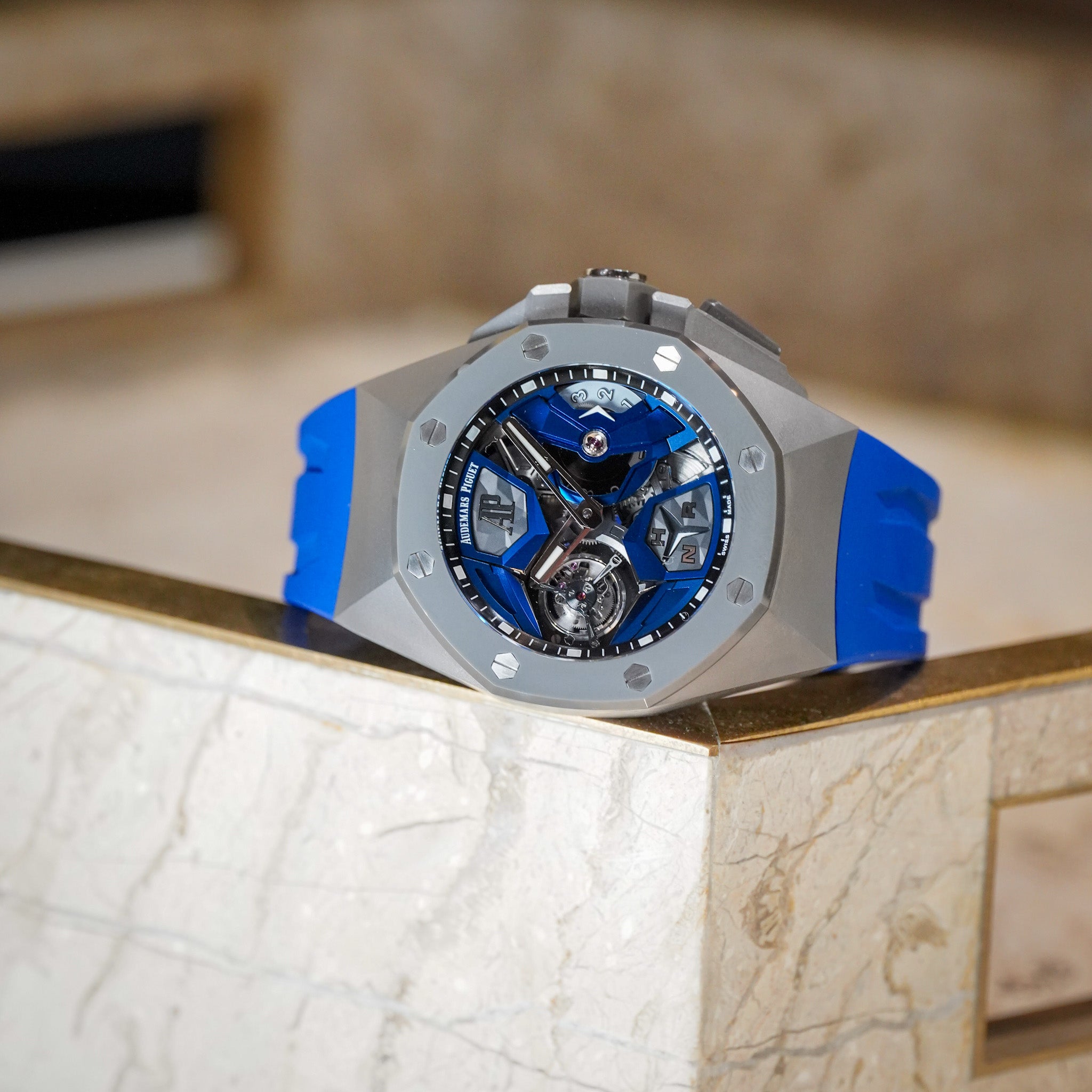 A luxury wristwatch with a silver-toned octagonal bezel, a blue and silver skeleton dial, and a bright blue rubber strap, resting on a marble ledge.