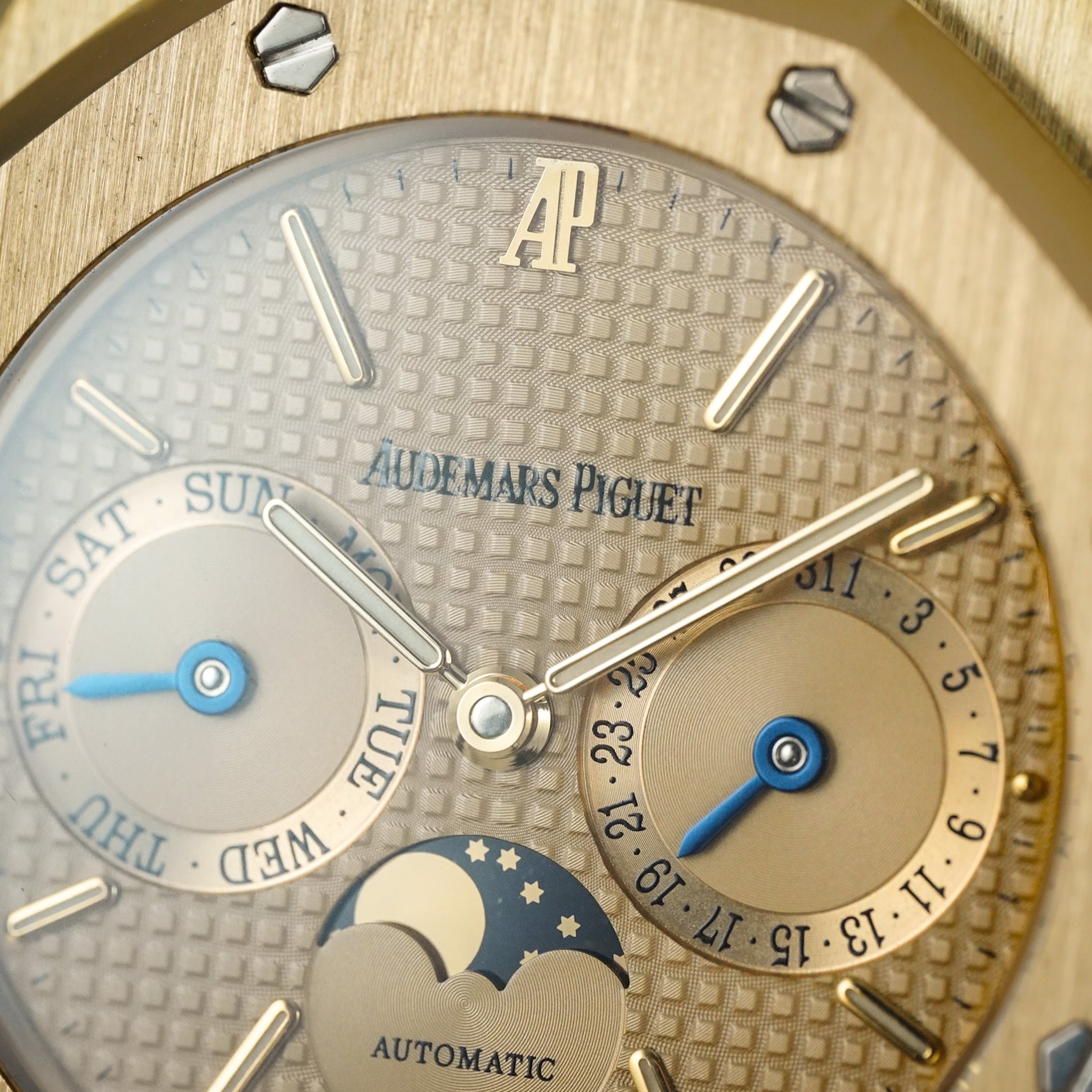 Close-up shot of a gold Audemars Piguet watch dial showing the AP logo, and three subdials for the day, date, and moonphase.